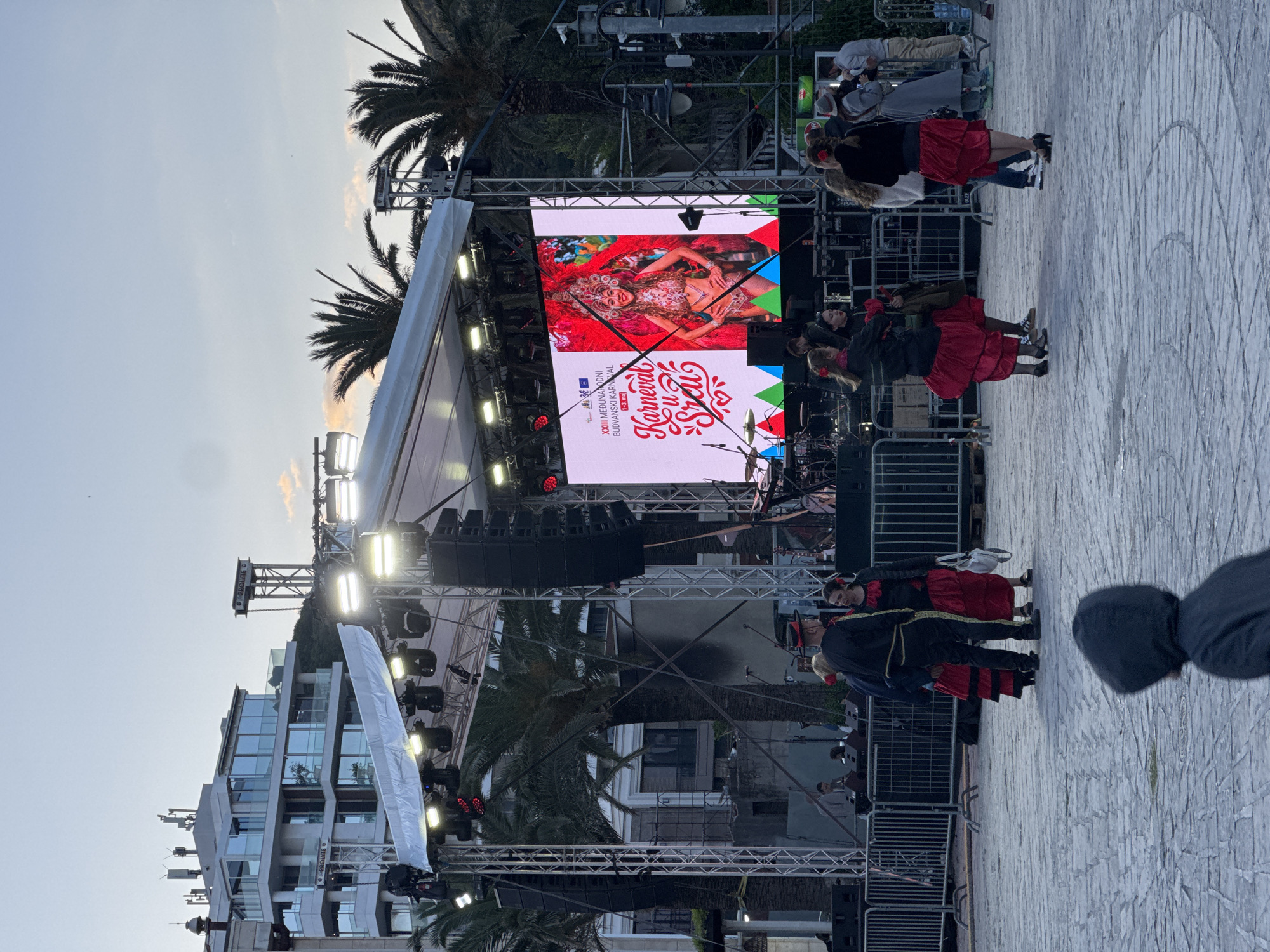 The image captures a scene in Budva, Montenegro, showcasing a stage setup outdoors. A large screen displays promotional imagery for the XXII International Budva Carnival. Several people are present, some dressed in traditional attire featuring red and black colors. A stage structure, palm trees, buildings, and the sky, are also visible. The stage has lighting equipment and speakers arranged above and around the central screen. The environment includes buildings and palm trees suggesting an urban area within a tourist destination. The foreground reveals a section of the ground, potentially a plaza or street area made of paved stone.