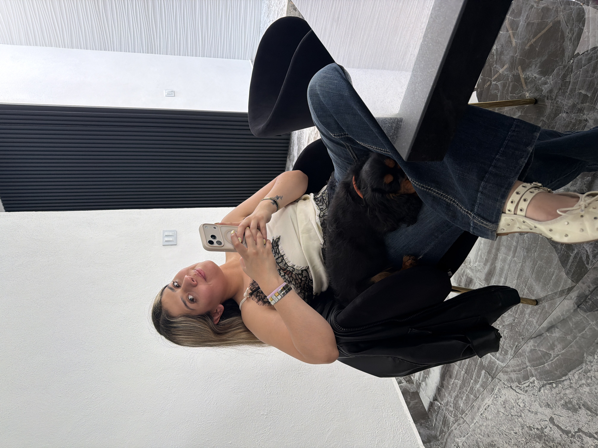 An indoor, medium shot captures a woman and a small dog in a modern setting in Puebla, Mexico. The woman, with light brown and blonde highlights in her hair, is seated on a black chair at a dark table with a grey, marble-like surface. She is wearing a white sleeveless top with black lace trim, blue jeans, and white studded flat shoes. Her left wrist is adorned with multiple bracelets. She holds a light-colored smartphone with a three-lens camera in both hands, directing her gaze towards the camera with a calm expression.
Resting comfortably on her lap is a small, dark-furred dog with tan markings, appearing to be asleep or at rest. The background features a white wall with a subtle textured pattern, a section of wall with horizontal dark panels, and a simple electrical outlet or switch. The overall lighting suggests it is daytime. No explicit text is discernible within the image.