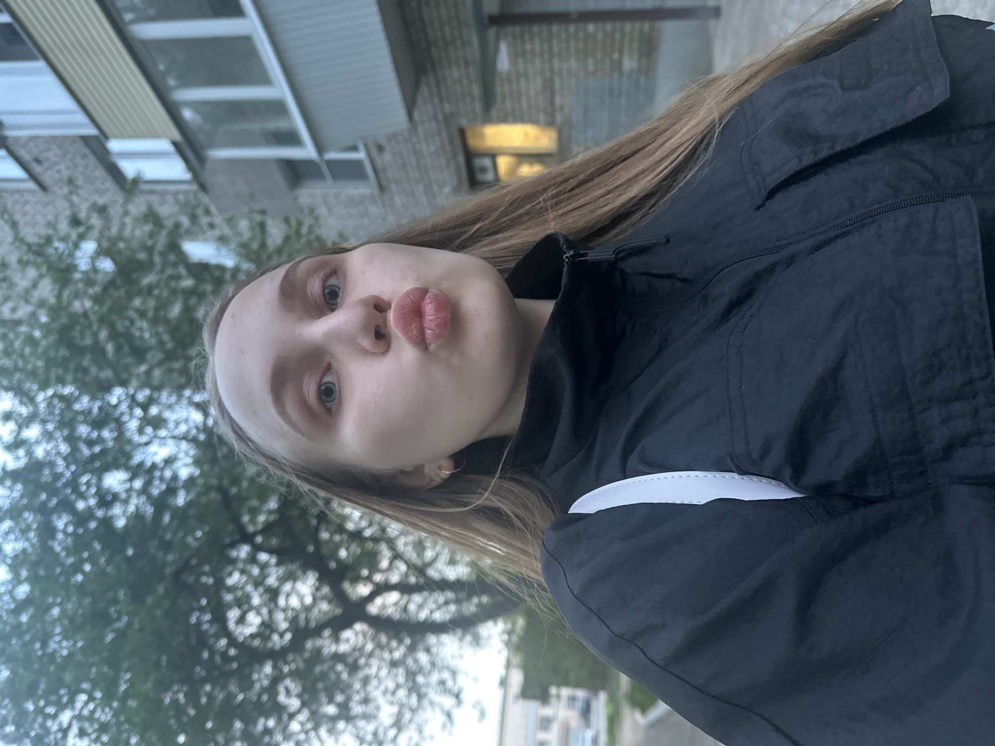 A young woman poses for a selfie outdoors, with a neutral expression and pursed lips. She is wearing a black jacket with a white stripe on the sleeve. Her long, light brown hair is parted to the side. Behind her, the background includes a multi-story building with windows, a tree with green leaves, and part of a street. The scene appears to be in a residential area of Chaikovsky, Russia.