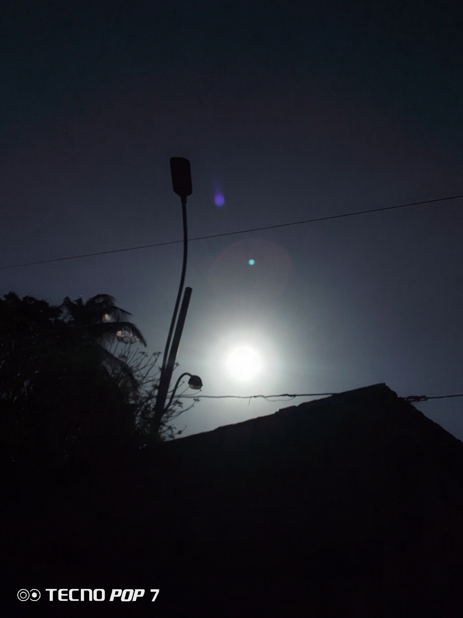 The image depicts a low-angle view of a night scene in Altagracia, Venezuela. The main subject is a bright light source, possibly the moon or a street lamp, positioned in the center of the frame. The scene is dominated by silhouettes of various objects against the dark sky. A street lamp is visible on the left side, with a bent pole and a downward-facing light fixture. A building roofline cuts across the lower part of the frame. A palm tree and other foliage are also visible on the left, rendered in dark silhouette. Power lines stretch horizontally across the upper portion of the image. The lower left corner displays the text "TECNO POP 7".