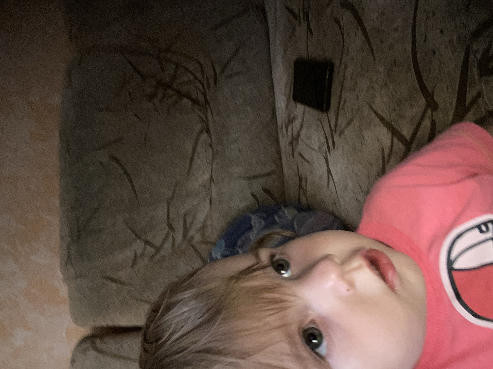 A close-up, eye-level, indoor portrait shows a young child with blonde hair and green eyes wearing a pink shirt. The child is looking to the left with a slightly parted mouth. Behind the child, a brown couch is visible, with a dark rectangular object, possibly a phone, resting on it. The background wall appears textured and light-colored.
