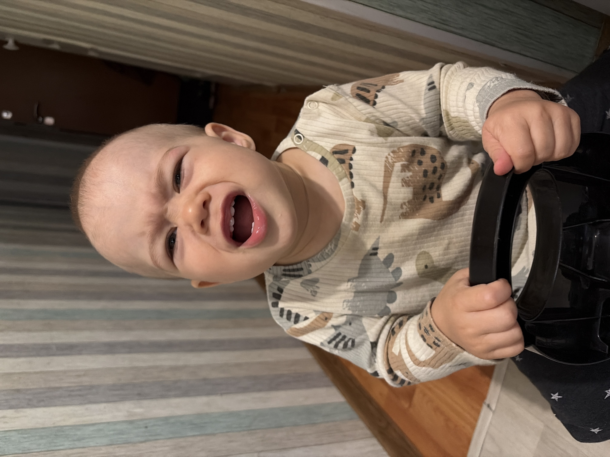 A baby with a bald head, wearing a long-sleeved shirt with animal prints, is crying and holding onto a black object that appears to be part of a toy or piece of equipment. The baby's mouth is open, and their face is contorted in a grimace. The background shows a striped surface, possibly a wall, and a wooden floor. The photo was presumably taken indoors. There is no discernible information in the photo that places it specifically in Chaikovsky, Russia.