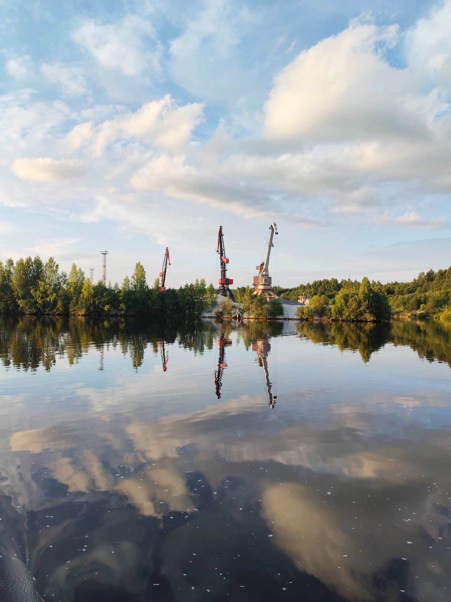 The image depicts a scenic view of a body of water reflecting the sky and surrounding trees, with several industrial cranes visible on the far bank. The water is calm, creating a mirror-like surface that doubles the image of the sky and the lush green trees lining the shore. Three prominent cranes, two red and one beige, stand tall against the horizon. The red cranes are positioned closer together, while the beige crane is slightly further to the right. The sky is adorned with fluffy white and grey clouds, suggesting a partly cloudy day. The scene appears to be a port or industrial area situated in a natural environment, possibly along a river or lake. Based on the provided context that this is in Aleksandrovskoe, Russia, it is likely a river port or an industrial zone on a body of water within or near the settlement. The image captures a peaceful yet industrious landscape, showcasing the coexistence of nature and human infrastructure.