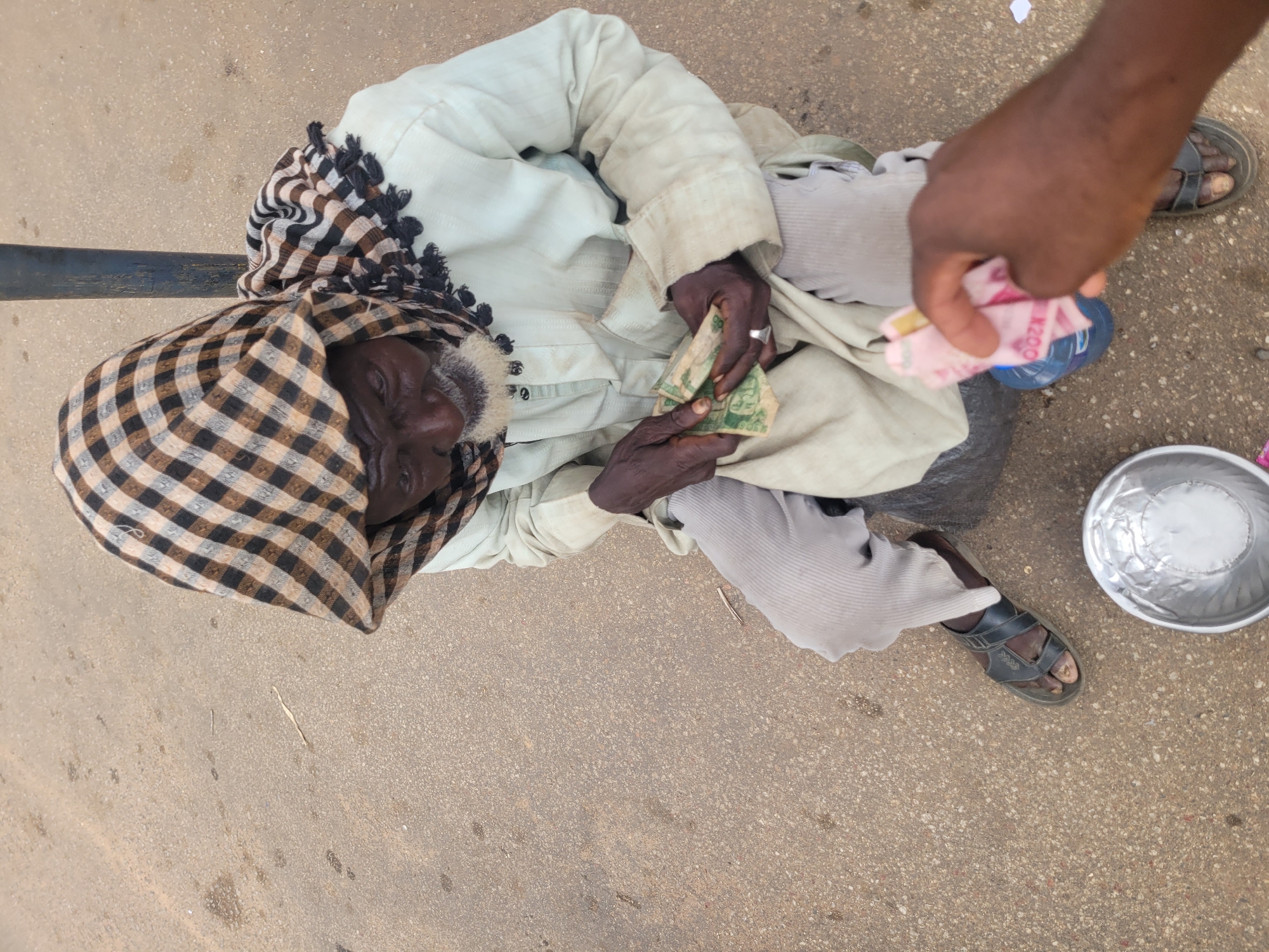 An overhead shot shows an elderly African man, possibly begging, sitting on the ground outdoors in Jos, Nigeria. He is wearing a light-colored robe and a checkered headscarf, holding a small wad of folded bills in his hands. In front of him is a silver bowl, and a hand extends into the frame from the right, offering a pink bill. The ground appears to be paved or asphalt, with scattered debris.