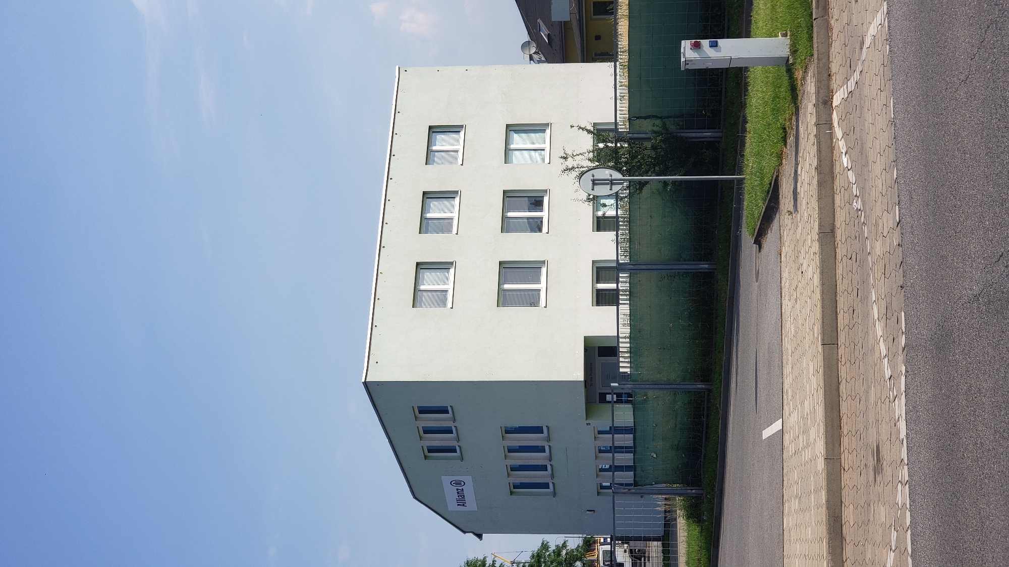 The image shows a two-story light-colored building with numerous windows. A green fence runs along the front of the property. The building has the "Allianz" logo on the left side. In the foreground, there is a paved area with road markings, likely a parking lot or driveway. The sky is clear blue. The building is located in Prešov, Slovakia.