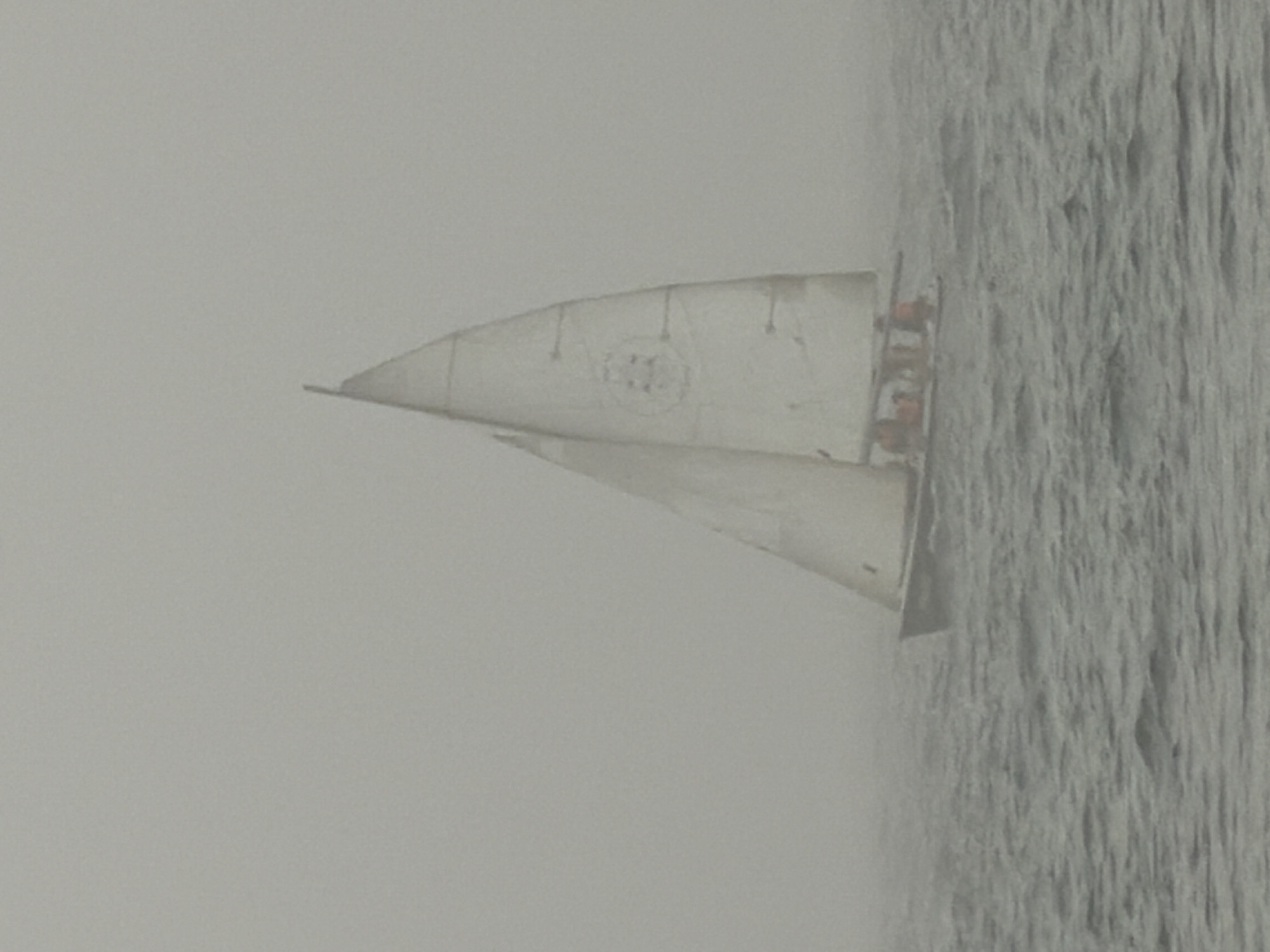 The image shows a sailboat navigating through dense fog or mist, seen from a distance on choppy, grey water. The background is an expansive, featureless light grey, indicating extremely low visibility.
The sailboat has two white sails unfurled. On the main sail, a circular emblem is visible, which appears to contain the number "11" or a similar symbol, though it is indistinct due to the low resolution and atmospheric haze. Several small, dark figures, presumably people, are visible in the cockpit area of the boat, suggesting an active sailing scenario.
The scene conveys a sense of isolation or challenging navigation due to the thick fog. The diffuse lighting prevents a clear determination of the time of day, but it is clearly daylight. The water surface shows texture, suggesting movement. While the image itself lacks explicit geographical cues, the provided context places the location in Tel-Aviv, Israel. No other distinct text or specific notable details are discernable apart from the sail marking.