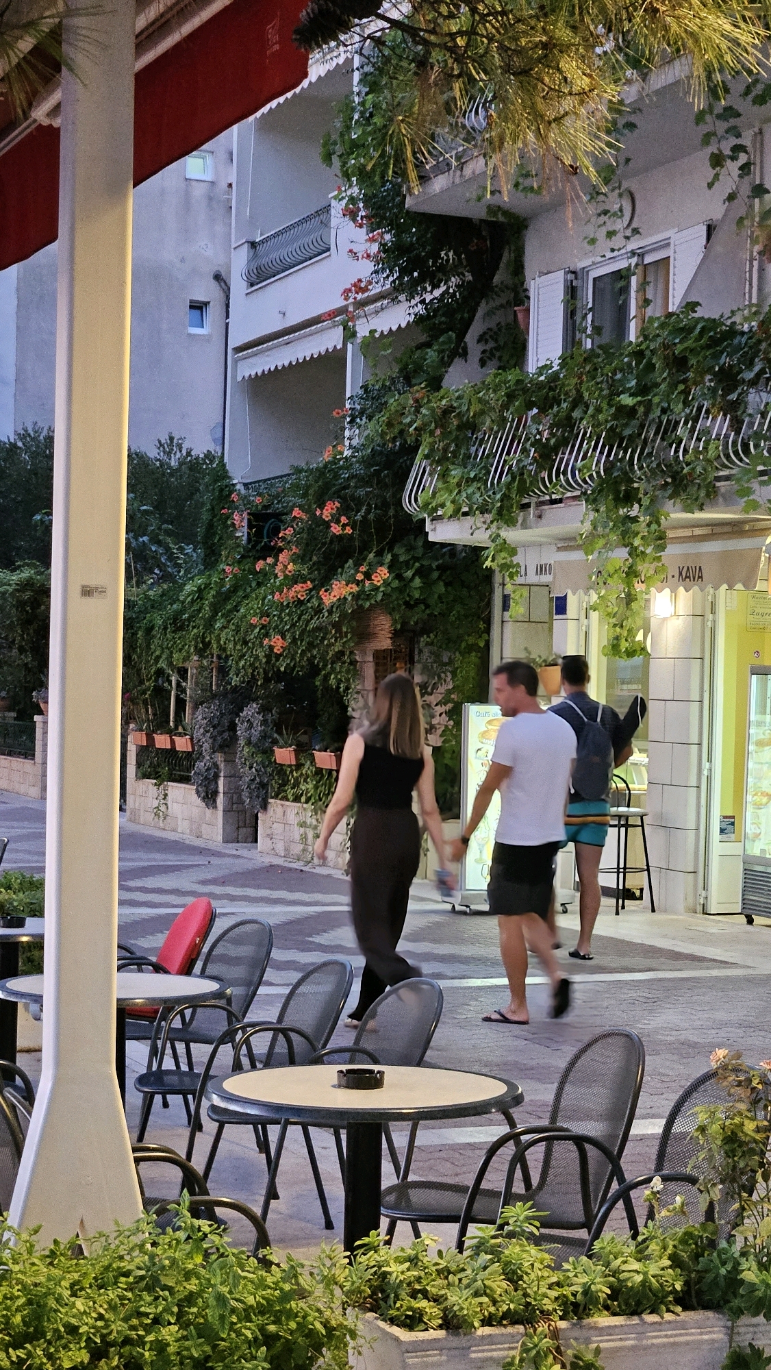 In Općina Tučepi, Croatia, a view captures an outdoor seating area with tables and chairs adjacent to a building with balconies adorned with greenery and flowers. A white column is prominent in the foreground. Three individuals are walking on a paved walkway in front of the building. A woman in a black sleeveless top and dark pants walks away from the camera, holding hands with a man wearing a white t-shirt and dark shorts. Behind them, another man with a backpack and blue shorts walks away from the camera. The building features windows and shutters, with some signage visible on the storefront. An ice cream display cooler is positioned to the right of the storefront. Lush vegetation lines the bottom of the frame and cascades down the building's exterior.