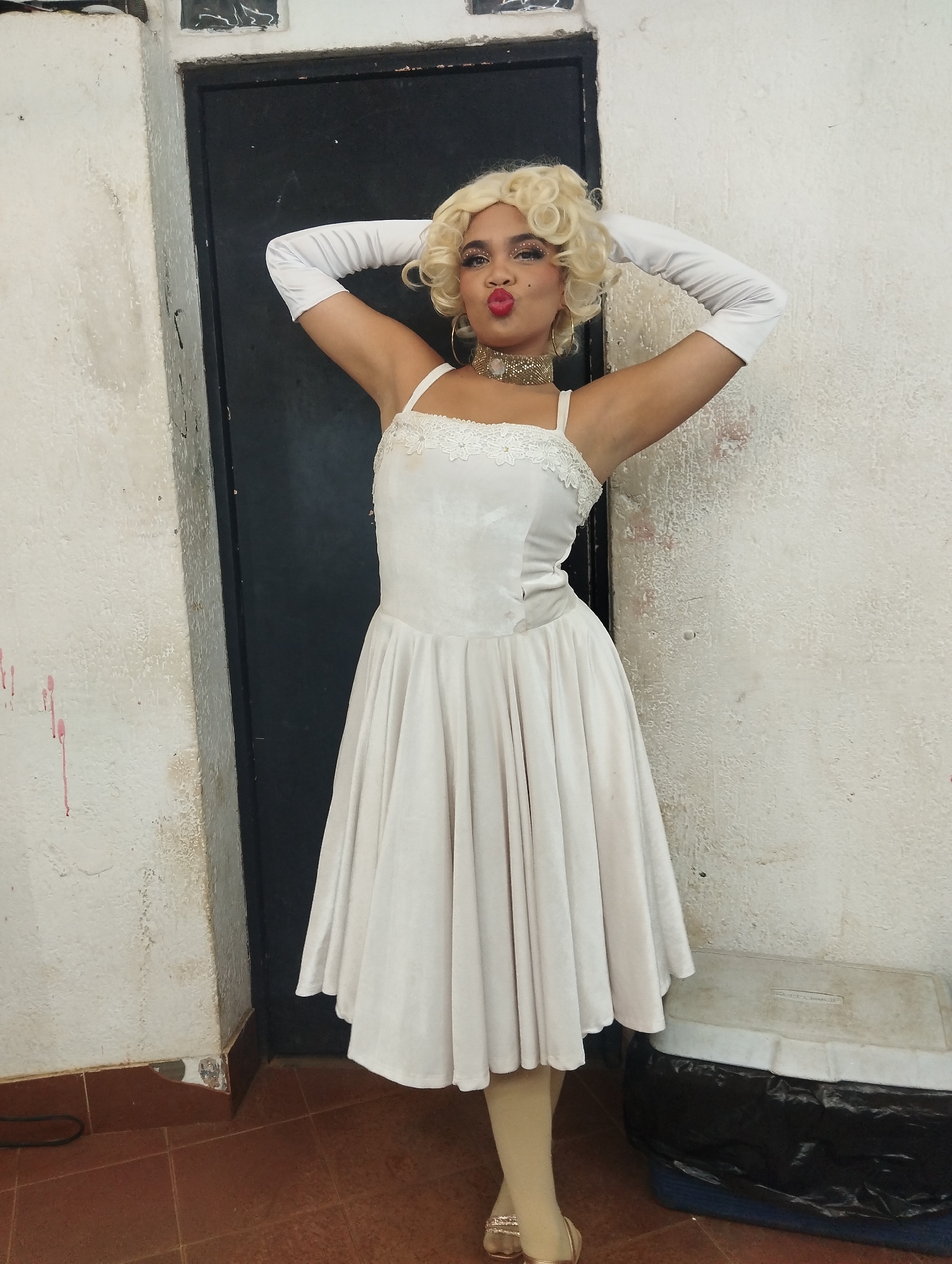 A person dressed as Marilyn Monroe poses in an indoor setting. The individual has fair skin, a blonde curly wig, red lipstick, and white gloves. They are wearing a white dress with spaghetti straps and a lace trim. The person is striking a pose, with their hands behind their head and a pouting expression. The location appears to be a backstage or dressing room area, with a dark door and aged white walls. A container with black trash bag beneath it is to the right of the person. The flooring is tile. The scene is not specific enough to precisely determine a location in Porlamar, Venezuela.