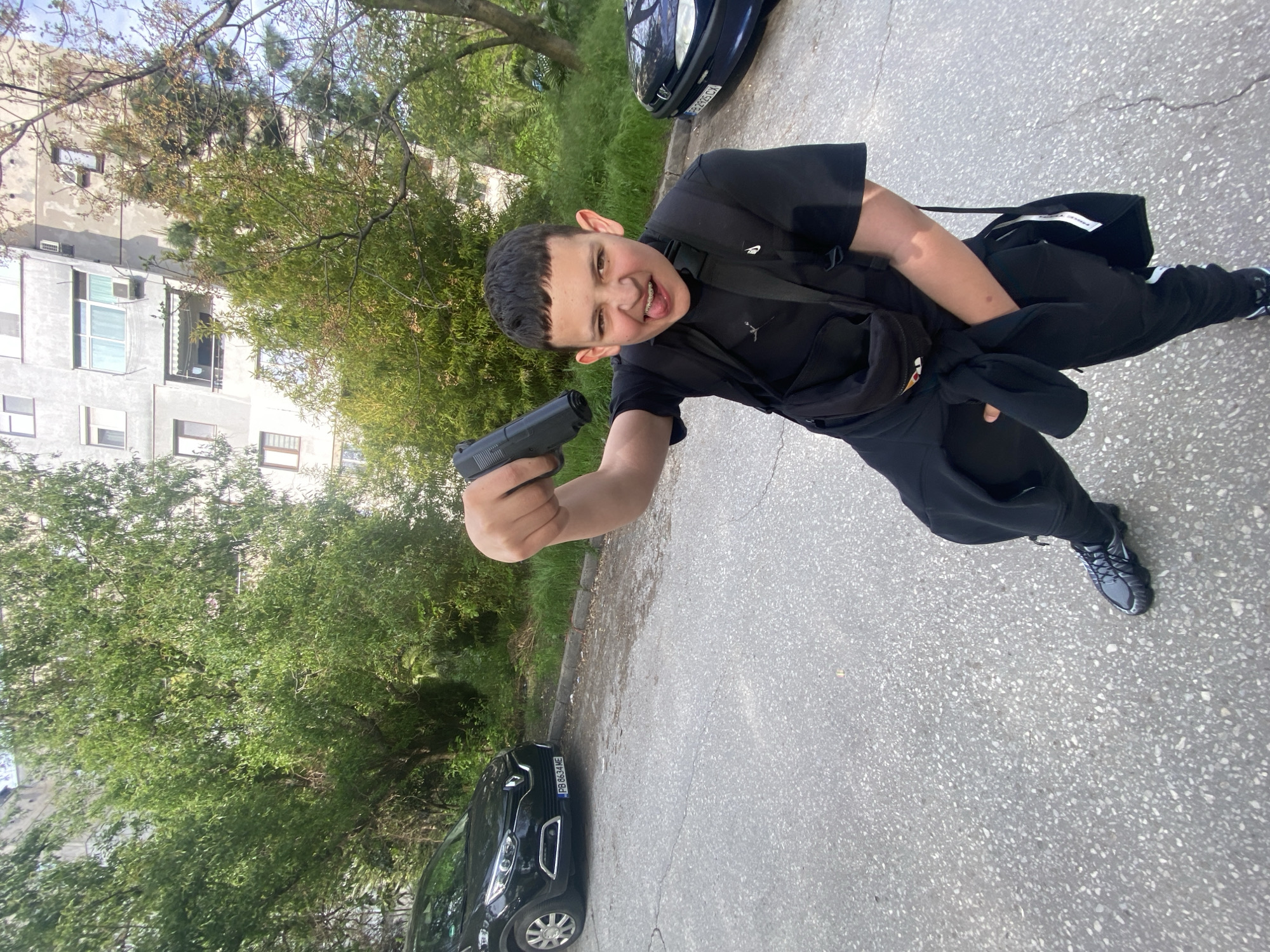 A youth stands on a paved surface with a handgun in his extended right hand. He is wearing black clothing, including a t-shirt, sweatpants, and sneakers. A black bag hangs across his chest. His facial expression appears to be a grimace or a playful aggressive expression, and he has braces. A car is visible behind him to his right and a building with trees to his left. The car license plate begins with "PB" suggesting that the location is Plovdiv, Bulgaria.