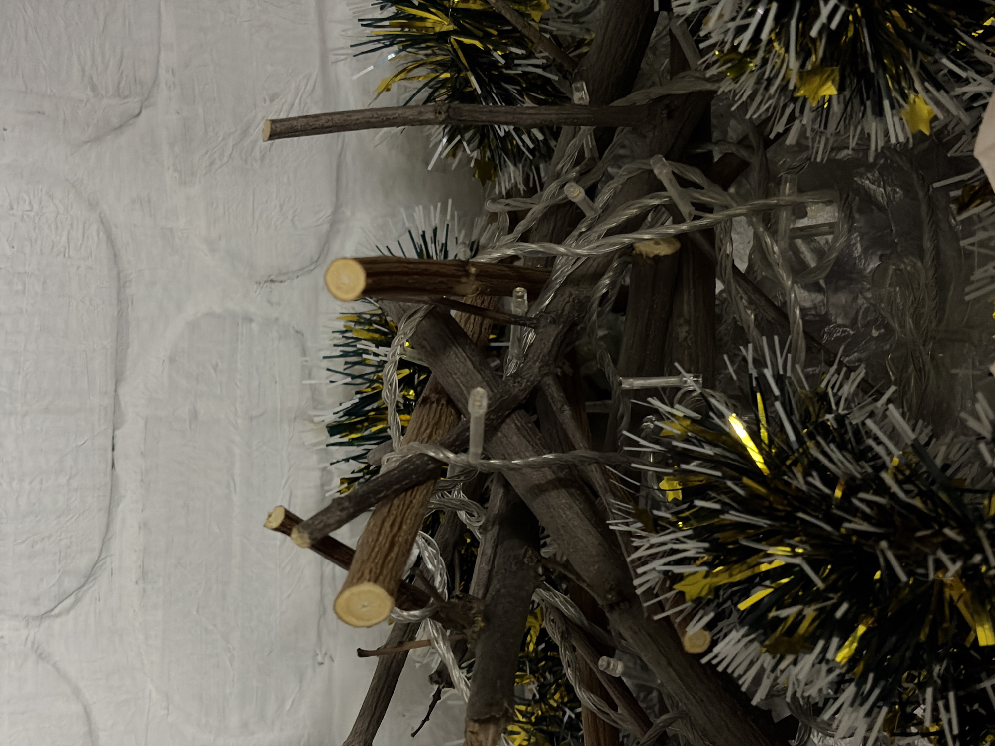 The image shows a Christmas tree made of sticks and twigs, wrapped with string lights and tinsel in black, white, and gold. The tree is placed against a white brick wall. There is no indication of a location within Port-Katon, Russia, or any people in the scene. The only objects are the Christmas tree, string lights, tinsel, and brick wall.