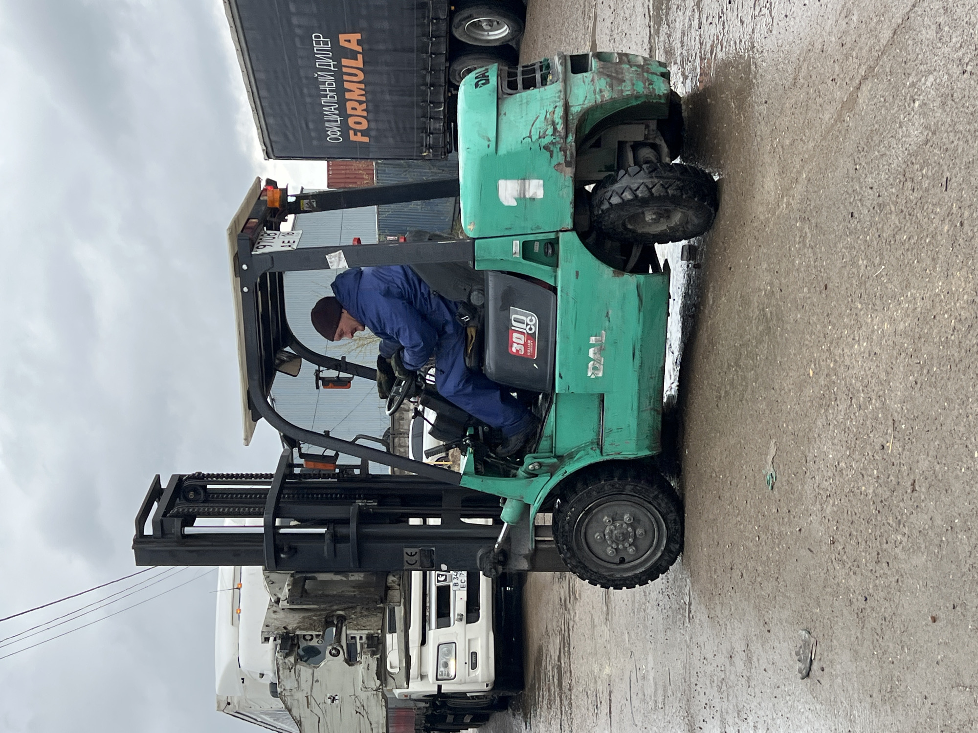 A man in blue work overalls and a dark beanie is seated in the operator's seat of a green forklift, which is positioned on a wet asphalt or concrete surface in an industrial yard. The man appears to be operating or preparing to operate the forklift, which has its forks partially raised.
The scene takes place outdoors under an overcast sky, with the damp ground suggesting recent rain or generally wet conditions. In the background, a large dark semi-trailer truck is parked, prominently featuring white text that reads "ОФИЦИАЛЬНЫЙ ДИЛЕР" (OFFICIAL DEALER) and "FORMULA". Another white semi-trailer truck is partially visible further in the background, displaying a license plate with "B 343 EC 17". The green forklift itself is well-used, showing signs of wear and tear, and has the numbers "3000" and "CC" visible on its engine cover, likely indicating its lifting capacity in kilograms. A large white number "1" is painted on the side of the forklift's body. A partial license plate "9706 AE 85" is also visible on the overhead guard of the forklift. The overall setting is an active industrial or logistics area during daytime.