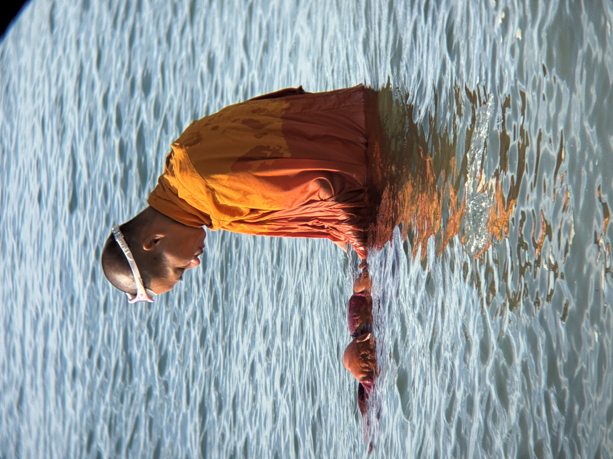In the city of Nha Trang, Vietnam, a Buddhist monk wearing orange robes and swimming goggles is in the water. He is looking down towards a submerged individual, also wearing a red garment. The water is calm with ripples, and the monk's reflection is visible. The scene takes place in a body of water, likely the ocean, given the location.