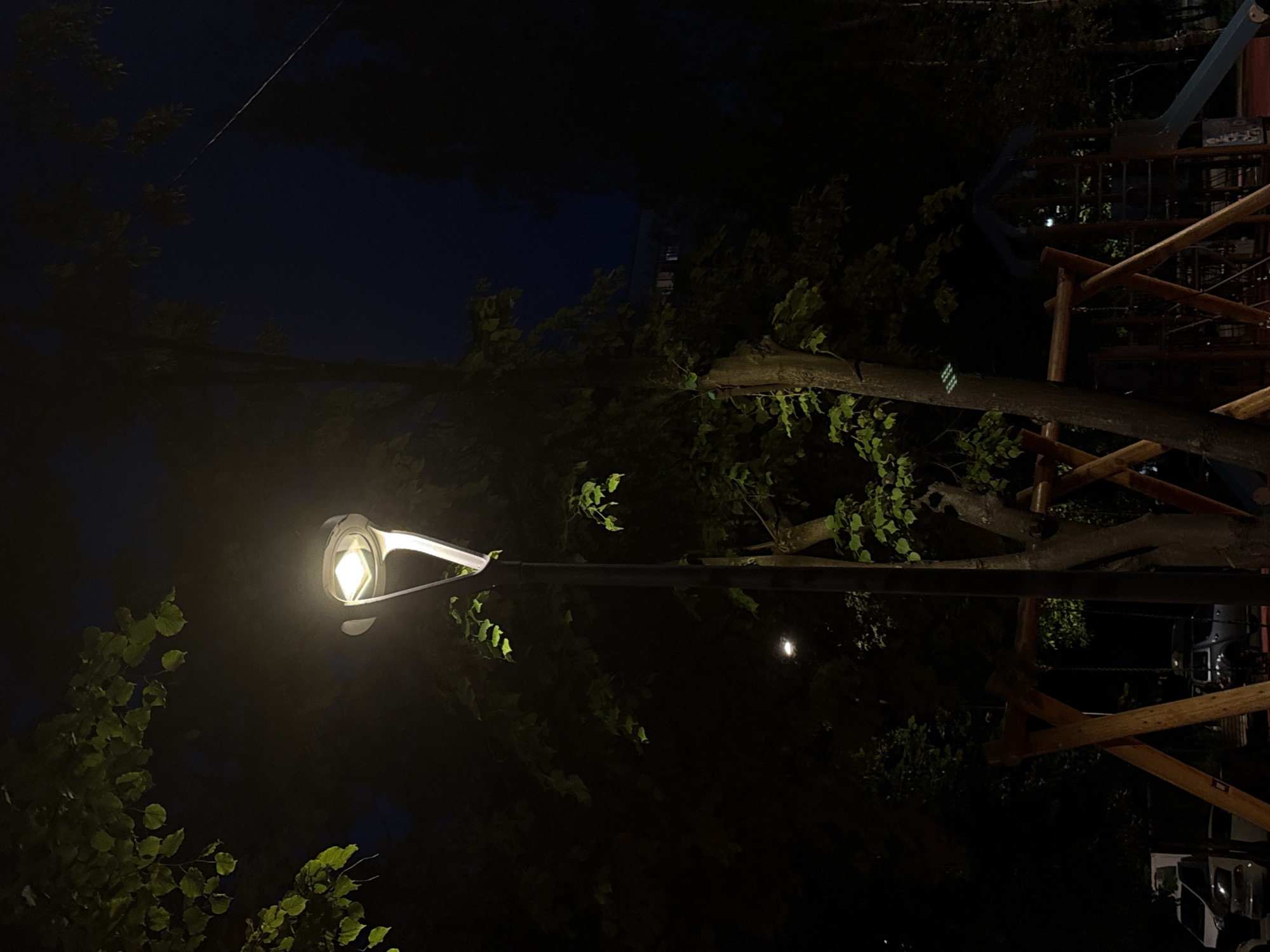 The image captures an outdoor nocturnal scene, likely a park or playground area in Moscow, Russia. A prominent, modern street lamp stands centrally, acting as the primary light source. The lamp's fixture is a light-colored, possibly metallic, structure shaped like an inverted 'Y' or 'V', emitting a bright, cool white light from a rectangular LED element.
Dense deciduous trees with dark trunks and branches frame the scene, with some green leaves illuminated by the street lamp, particularly in the upper left and mid-right sections. In the foreground and mid-ground, elements of a wooden playground structure are visible, including the distinct A-frame supports of what appears to be a swing set. To the far right, a portion of a blue or gray playground slide is discernible.
The sky is a deep, dark blue-black, characteristic of night. In the distant background to the left, outlines of parked vehicles, including a white van and a dark car, are faintly visible, suggesting proximity to a road or parking area. A subtle, secondary light source is present further back in the mid-left. A thin, dark line or wire runs diagonally across the upper right quadrant of the image. No people are present.