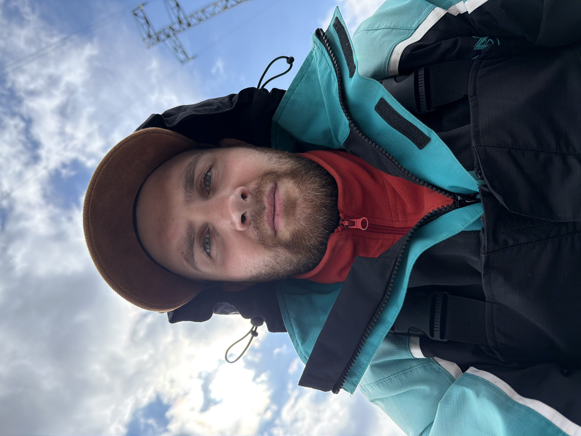 A man with a brown hat and beard is wearing a black, red, and teal jacket. He is outdoors under a cloudy blue sky with a power line structure visible in the background. The photo appears to be taken from a low angle, looking up at the man. The location is inferred to be Chaikovsky, Russia.