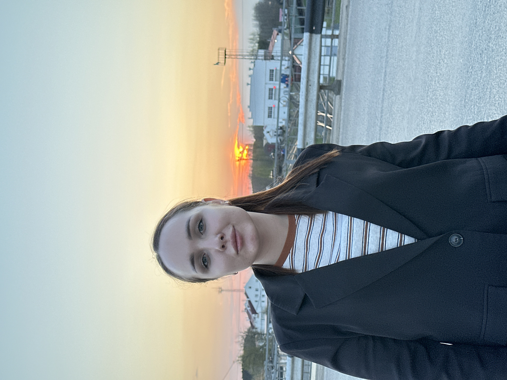 A photograph shows a woman standing in what appears to be the city of Chaikovsky, Russia, based on architectural style and the sunset's color, typical of that region. The woman is wearing a dark jacket over a striped shirt. The setting is outdoors, with the sun low on the horizon, creating a yellow and orange sky. Structures resembling buildings and utility poles are visible in the background.