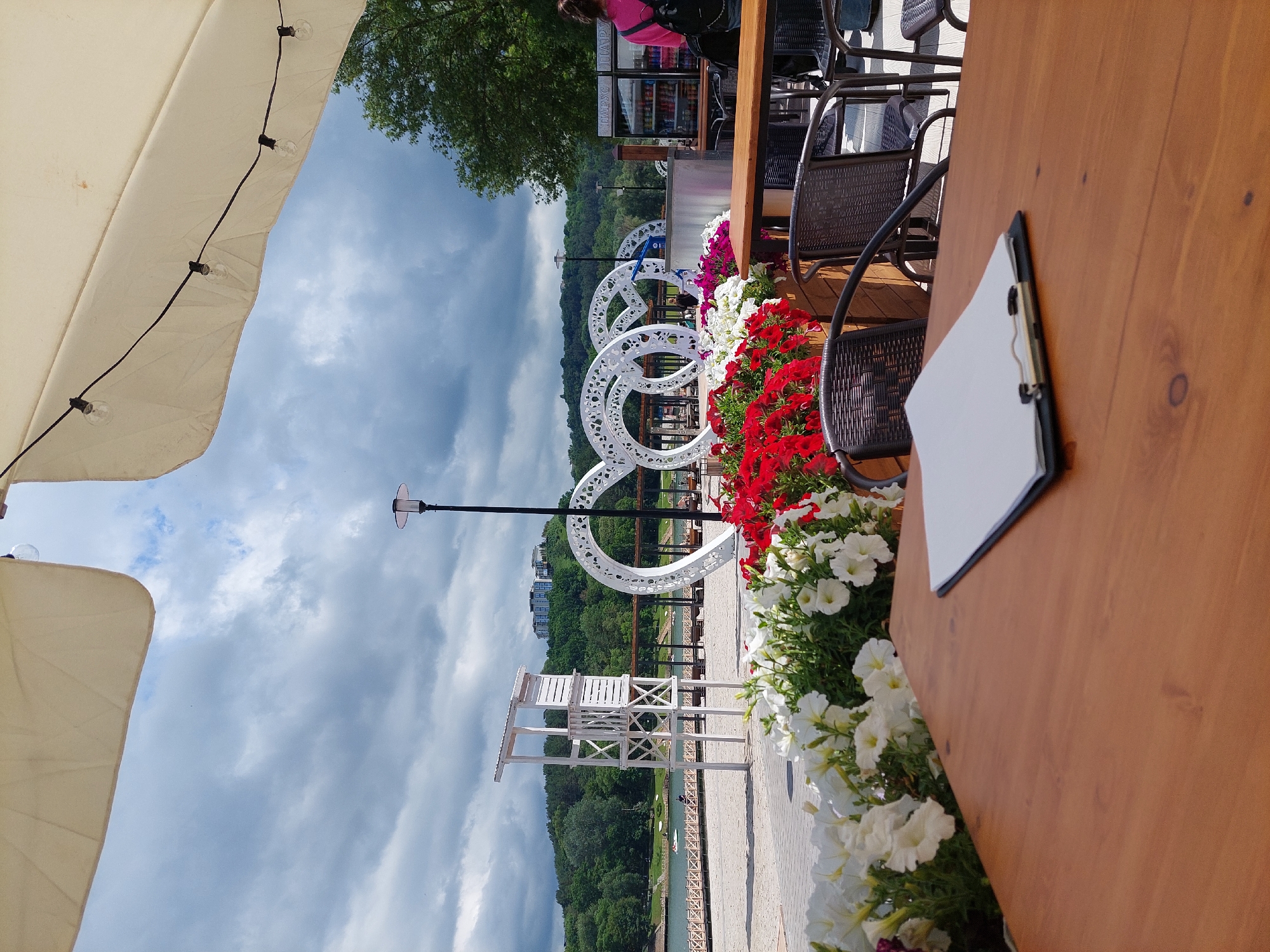 This image shows a view from an outdoor cafe table in Alikonovka, Russia, with a sandy beach and a lake in the background. A wooden table in the foreground holds a clipboard with blank white paper. Behind the table, there are several outdoor chairs, some of which are partially obscured by planters filled with white and red petunias. In the mid-ground, a white lifeguard tower stands on the beach. Further back, large white circular decorative structures are visible, along with a lake with a few boats and people. The background features a treed hill with buildings on top, and a cloudy sky. A person in a pink top and backpack is visible in the far right background.
