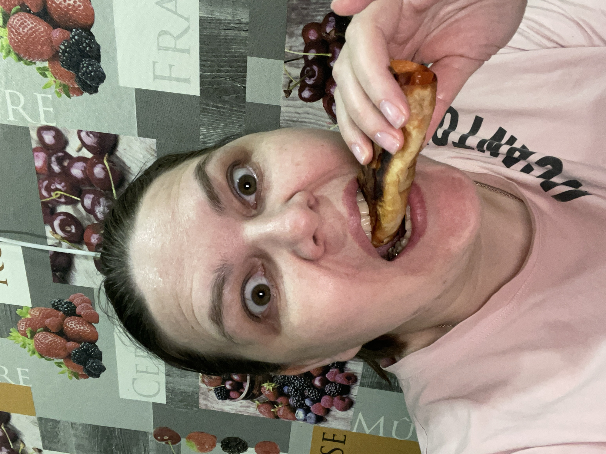 A close-up, rotated portrait captures a woman in a domestic setting, likely a kitchen or dining area. She is in the process of eating or about to bite into a golden-brown, cylindrical food item, resembling a fried pastry or roll, which she holds to her open mouth. Her eyes are wide and focused, conveying an intense, perhaps surprised or playful expression as she looks directly at the viewer.
She is wearing a light pink t-shirt with dark, block-lettered text visible across her chest, which appears inverted and partially obscured, possibly containing parts of words like "CHAMPION." Her hand, holding the food, shows naturally manicured nails with a clear or light polish. The background is a patterned surface, resembling a tablecloth or placemat, adorned with vibrant illustrations of various fruits, including strawberries, raspberries, blackberries, cherries, and blueberries. Fragments of text are also visible on this surface, such as "RE," "FRA," "CE," and "MU," suggesting words like "FRANCE." The image is oriented sideways, rotated 90 degrees clockwise. While the context provided is Noviy, Russia, there are no specific visual cues within the image itself to confirm this location.