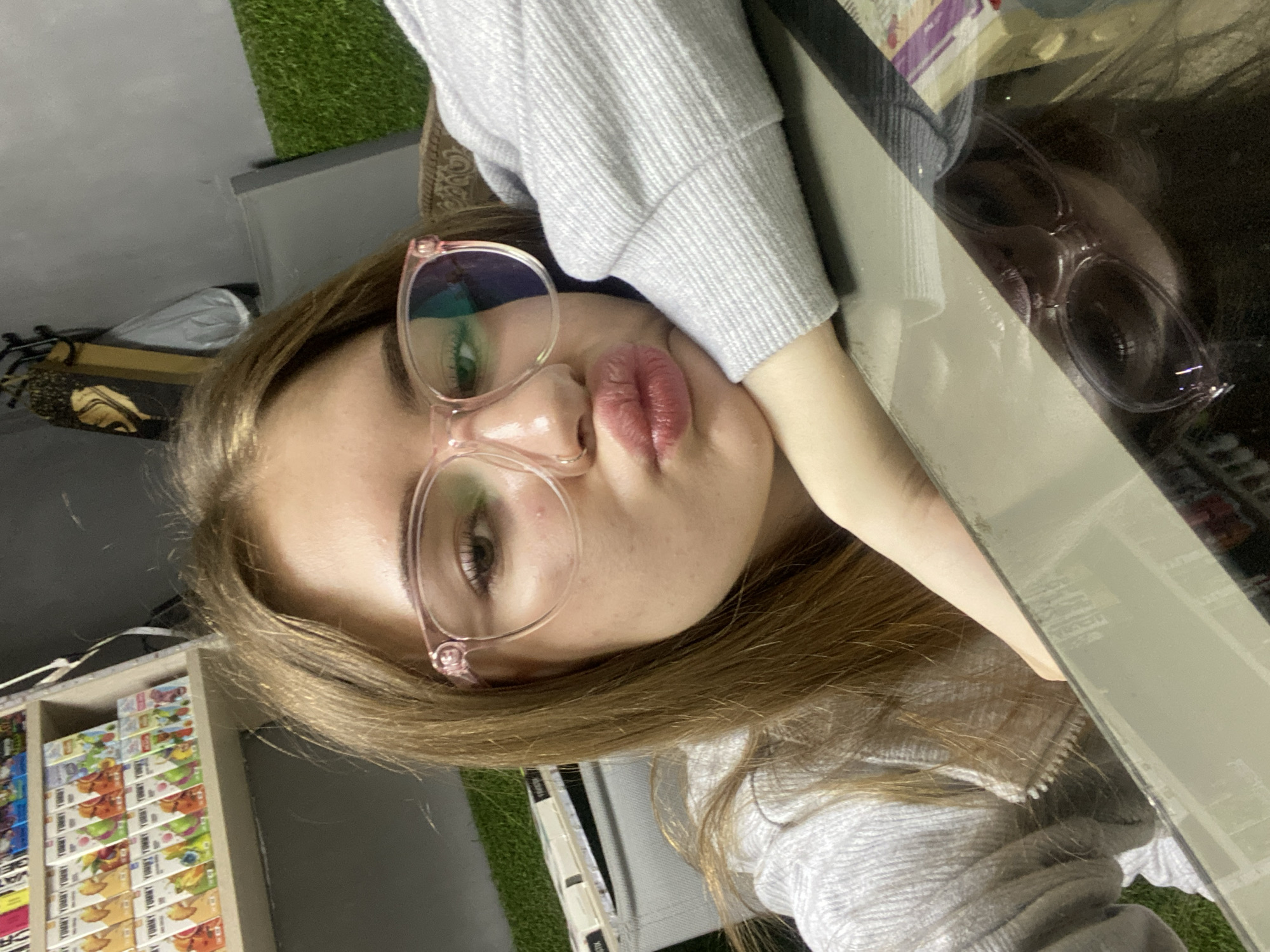 An indoor portrait captured in Дьяково, Russia, features a young female with long, light brown hair and pink-framed eyeglasses. She exhibits a small ring piercing in her left nostril and her lips are pursed in a pout. She is dressed in a light grey, textured, long-sleeved garment with a zipper detail. Her left arm is bent, and her chin rests upon her hand, which is positioned on a reflective, flat surface. This surface provides a dark, partial reflection of her face and glasses. The background reveals a multi-tiered shelving unit to the left, stocked with numerous small, uniformly packaged boxes bearing the label 'YUMMY TUMMY' and diverse colorful graphics. Adjacent to this, a grey wall transitions into an area featuring artificial green turf. Partially visible on the upper right wall are dark, decorative elements, and to the far right, a segment of a brown, textured furniture piece.