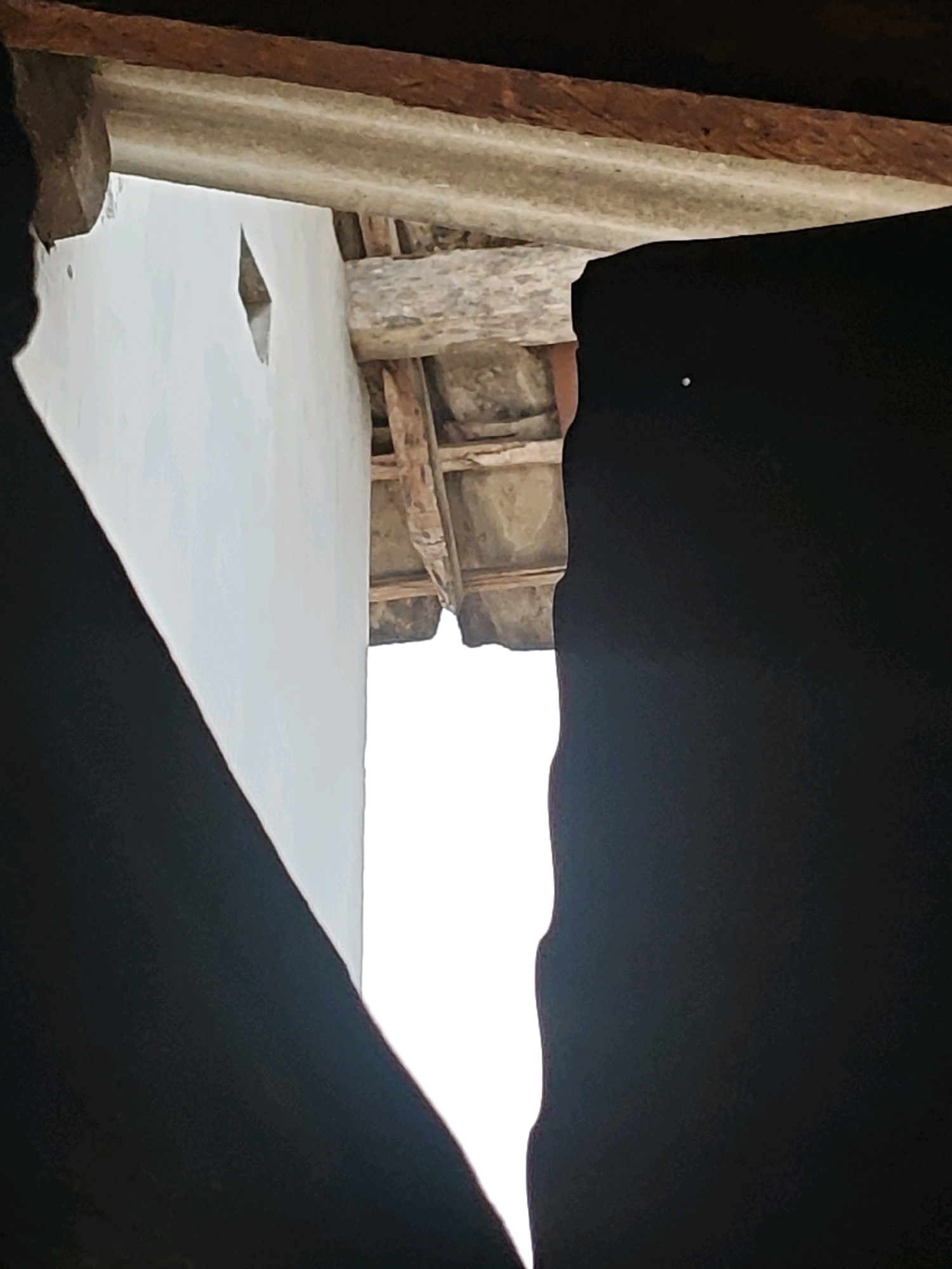 The image depicts an upward view through an opening. In the foreground, a dark, silhouetted object, possibly fabric or a shadow, frames the left and right sides of the scene, with its edges unevenly cut. Beyond this framing, a white wall occupies the left portion of the image, featuring a small, diamond-shaped opening. To the right of the wall, a rustic roof structure is visible, composed of wooden beams and what appears to be corrugated sheeting or tile. The background is predominantly a bright, blown-out white, suggesting a very bright sky or an overexposed area. The scene appears to be an exterior view looking upwards into a gap between buildings or structures. Given the Pagu, Indonesia location, this could be a view into a courtyard, alleyway, or between structures in a residential or traditional setting, common in areas with older architecture or informal settlements. There are no people or animals visible in the image, and no specific actions or interactions are captured. The overall impression is one of looking out from a sheltered or enclosed space into a brighter, open area.
