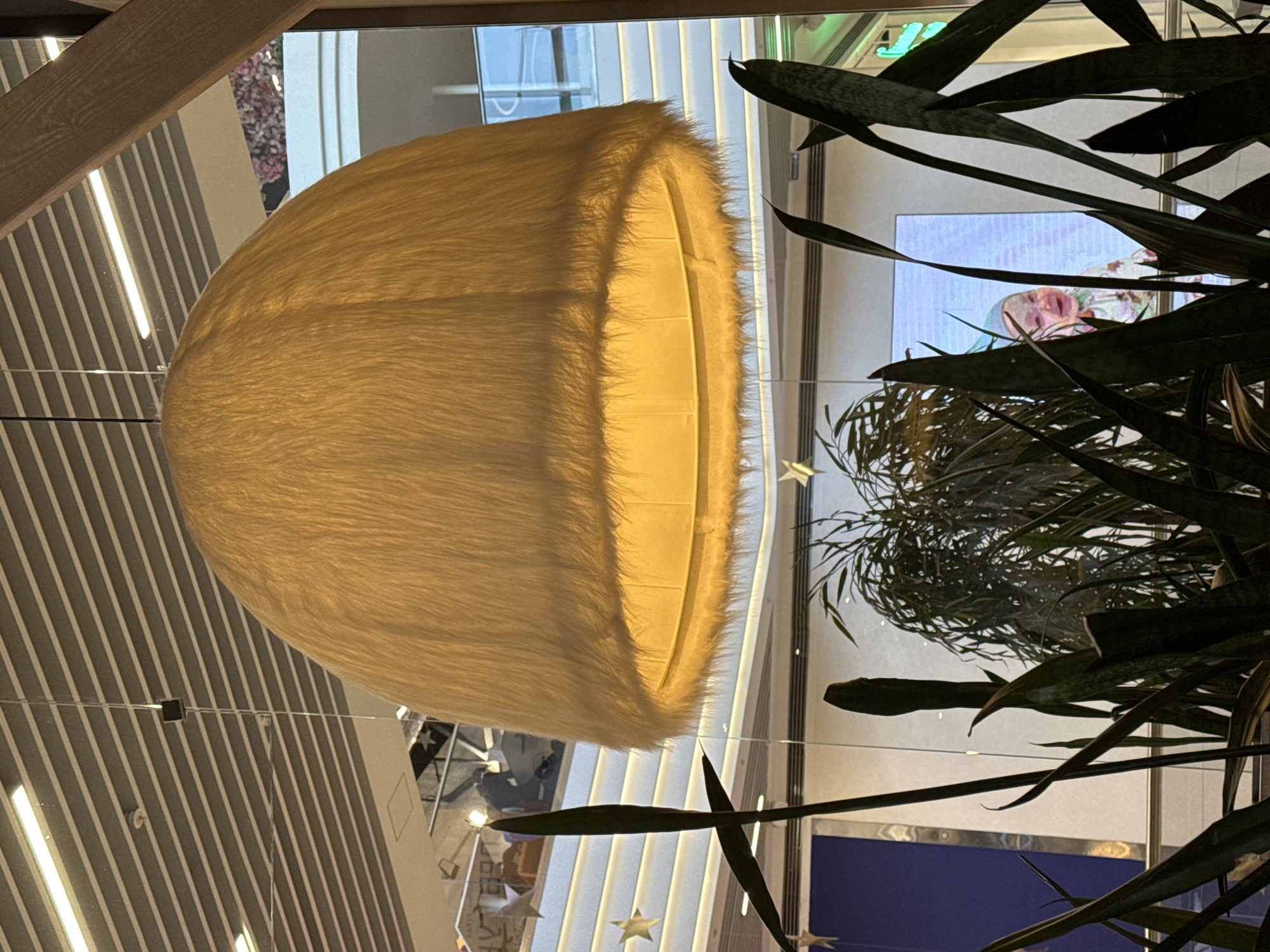 The image depicts an indoor scene within a multi-level commercial establishment in Perm, Russia. A large, illuminated, dome-shaped pendant lamp, covered in light yellow fur-like material, hangs centrally from a modern slatted ceiling equipped with linear lighting and a fire sprinkler. Dark green, sword-like leaves of Sansevieria plants dominate the immediate foreground, partially obscuring the background. Through the reflective glass panels in the background, a multi-story interior is visible, featuring glass railings, stepped surfaces, and additional star-shaped decorative elements. Reflected imagery includes a blurred individual pushing a floor cleaning machine on an upper level and a prominent digital display screen exhibiting a smiling infant wearing a light-colored cap. A second, larger green plant is partially discernible behind the foreground foliage.