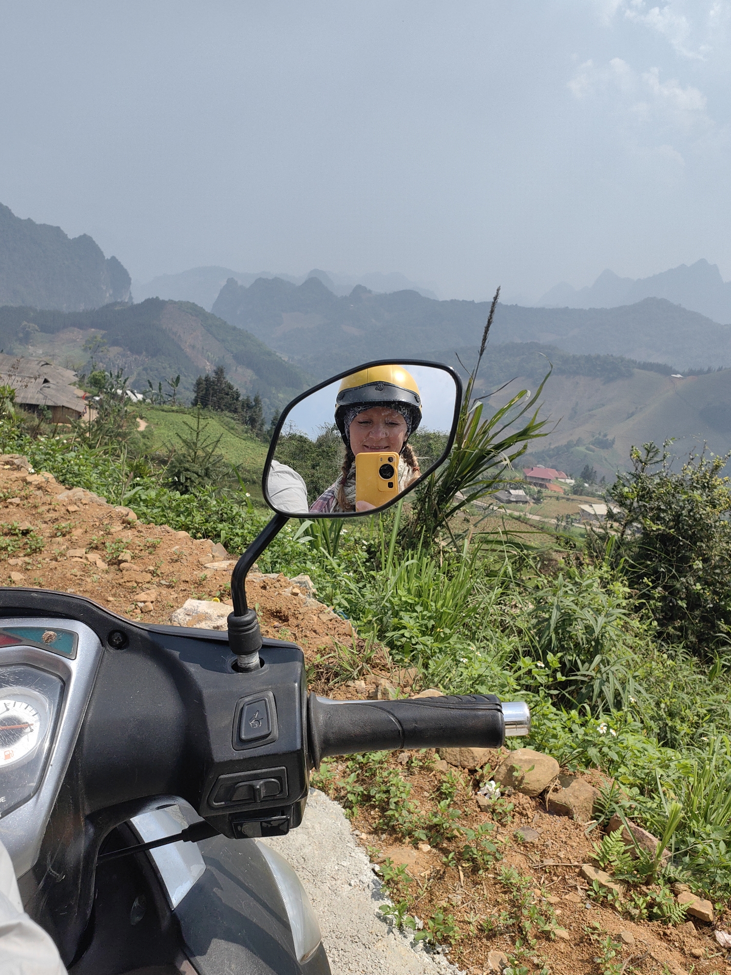 A woman in a yellow helmet takes a selfie in a motorcycle mirror overlooking a valley with tiered slopes and buildings. The motorcycle’s handlebars and front of the bike are visible in the foreground. The background features a hazy sky over green hills and mountains. The location is Xã Cần Yên, Vietnam.