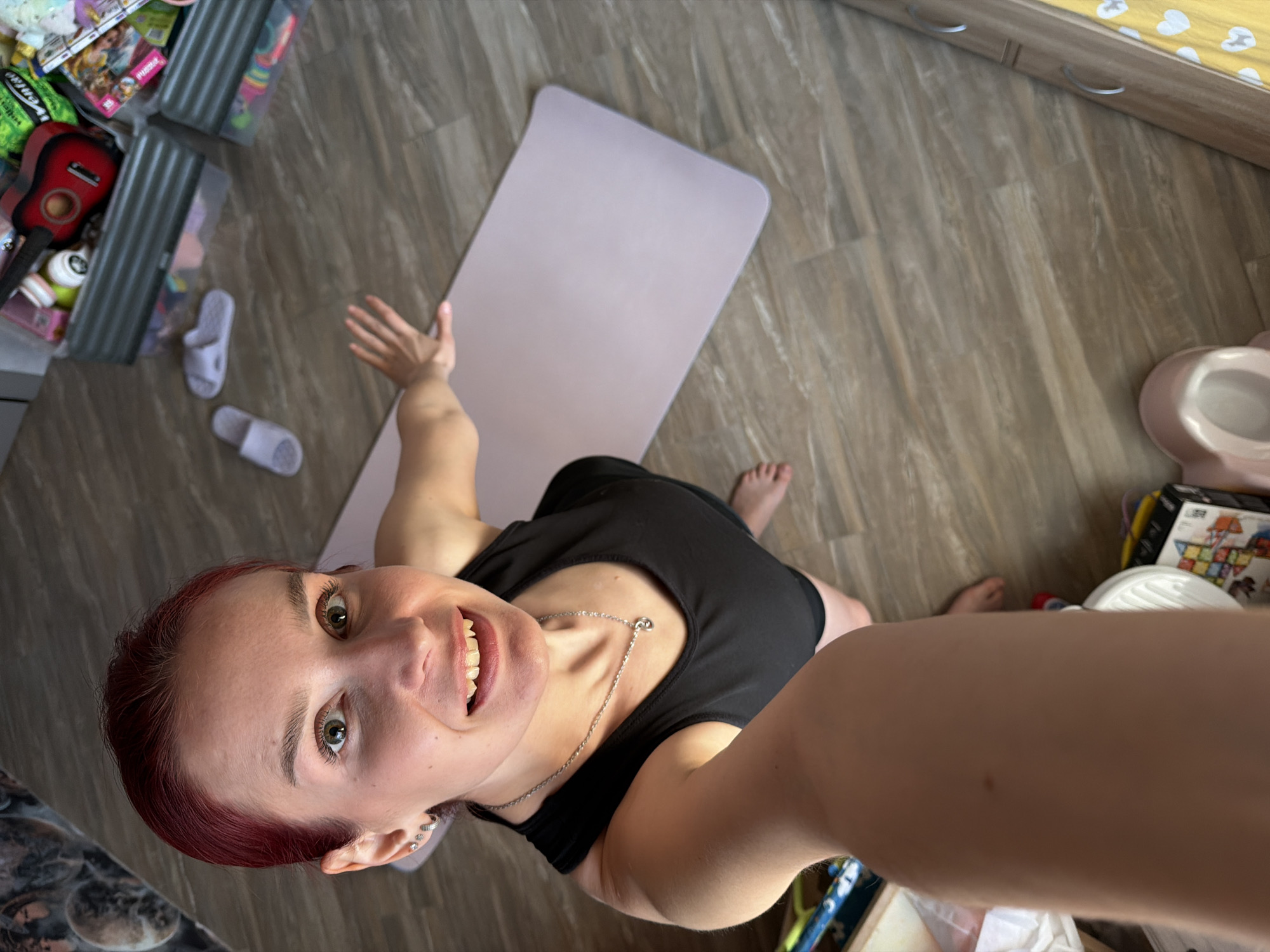A high-angle selfie captures a young adult woman with reddish-brown hair, dark green eyes, and a broad, cheerful smile, looking directly at the camera. She is wearing a black tank top and a silver necklace. Her right arm extends towards the camera, while her left arm is raised with an open hand gesture. She appears to be sitting or kneeling on a light brown wooden or laminate floor in an indoor setting in Chaikovsky, Russia.
Behind her is a light purple yoga mat. The surroundings suggest a domestic environment, possibly a child's room or a shared living space. To her left, various items are visible, including a red toy guitar, storage bins containing toys (one partially shows "VEMOVE" and another "PRINCESS 25 18 PCS"), and a pair of white slip-on sandals. To her right, a light pink child's potty trainer is on the floor, next to a black box with "ME" visible, possibly from a building toy set. A wooden cabinet with silver handles and yellow-and-white patterned bedding is visible in the upper right background. The bright natural lighting suggests it is daytime. The woman's joyful expression and the yoga mat imply she might be about to engage in or has just finished a fitness activity.