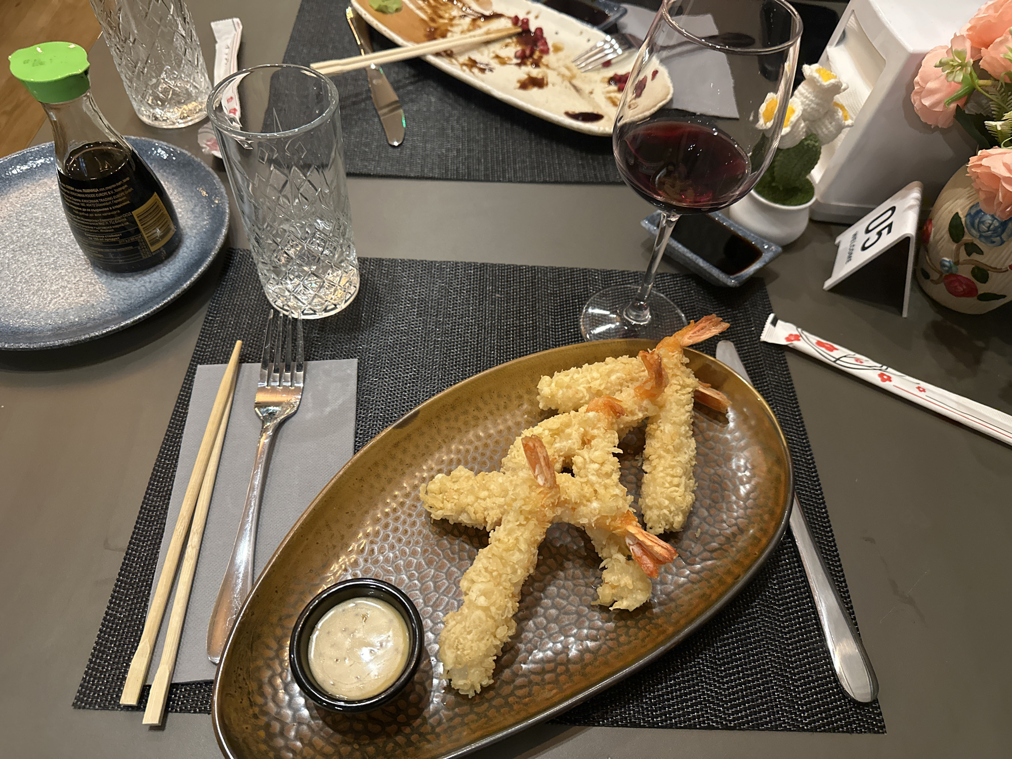 The image depicts a table setting at a restaurant, likely indoors. The table is covered with dark gray placemats and a solid dark gray tabletop. Food and beverages are arranged on the table.
In the foreground is a plate of fried shrimp tempura with a dipping sauce. Next to the plate are chopsticks and a fork. A wine glass filled with red wine is present, along with a small dish of soy sauce. A glass of water is also on the table.
In the background, there is another plate with remnants of a meal, including pomegranate seeds. Used chopsticks and cutlery are also present. A small soy sauce bottle with a green cap sits on a blue plate.
A small floral arrangement and a table number stand are also visible in the background.
The location cannot be verified from the image alone, but the prompt suggests it is a restaurant in Sofia, Bulgaria. No people are visible in the image. The focus is on the food and table setting.