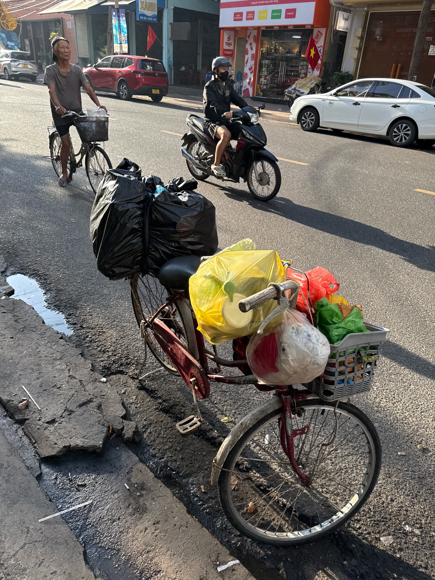 The image depicts a street scene in Nha Trang, Vietnam. In the foreground, a red bicycle is laden with numerous plastic bags filled with goods, primarily white, yellow, red, and green. These bags are secured to the bicycle's handlebars and a basket attached to the rear. A black plastic bag is also prominently displayed on the rear of the bicycle. The bicycle is parked on the right side of the road, adjacent to a damaged section of the asphalt. In the middle ground, a man wearing a helmet and a face mask rides a motorcycle. Behind him, a red car is visible, and further in the background, white cars and various storefronts line the street. The lighting suggests it is daytime, with strong shadows cast across the road. The environment appears to be an urban setting with typical street infrastructure.