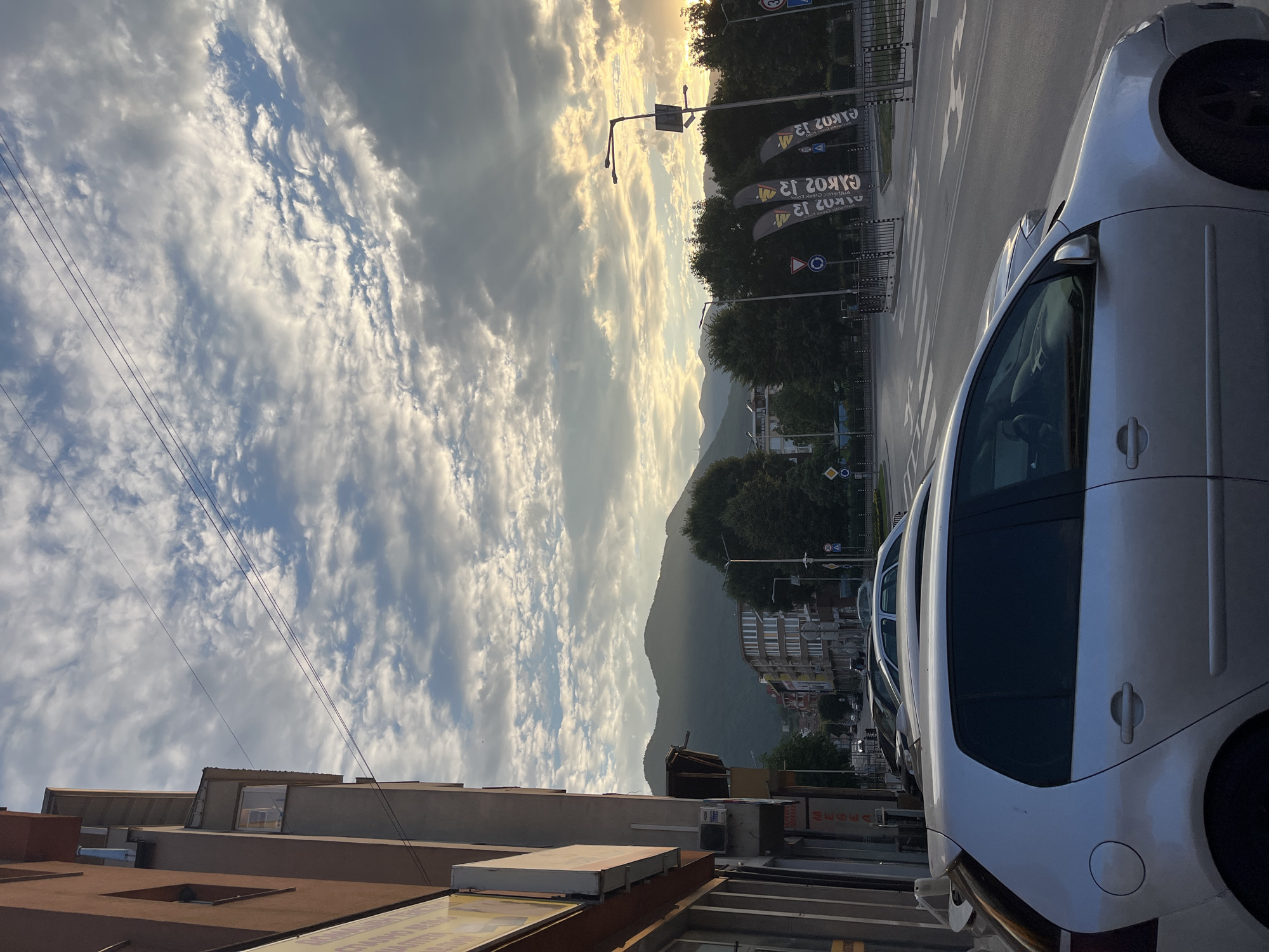 This is a vertical, slightly tilted, eye-level shot of a street scene in Gotse Delchev, Bulgaria, likely during late afternoon or early evening. The sky is filled with dramatic, sun-drenched clouds, suggesting fair weather with some atmospheric activity. The sun's warm glow filters through the clouds, illuminating the scene with a soft, golden light.
In the foreground, the silver side of a car is prominent, suggesting a parked vehicle. Beyond the car, a street stretches out, marked with white directional arrows. Several tall, dark banners are visible along the roadside, each displaying text. One banner clearly reads "GYROS 2012" in white lettering, with additional text below that is partially obscured but appears to say "Authentic Greek Food." Traffic signs, including a circular sign with a red border and a road symbol, are also present.
Further down the street, buildings line the road, appearing to be a mix of commercial and residential structures. Trees with lush green foliage are interspersed between the buildings and along the hillside in the distance. A prominent mountain range forms the backdrop, its slopes softened by the ambient light.
Overhead, several dark power lines crisscross the sky, a common urban feature. The overall impression is of a quiet, everyday scene in a town nestled near mountains, bathed in the beautiful light of a setting sun.