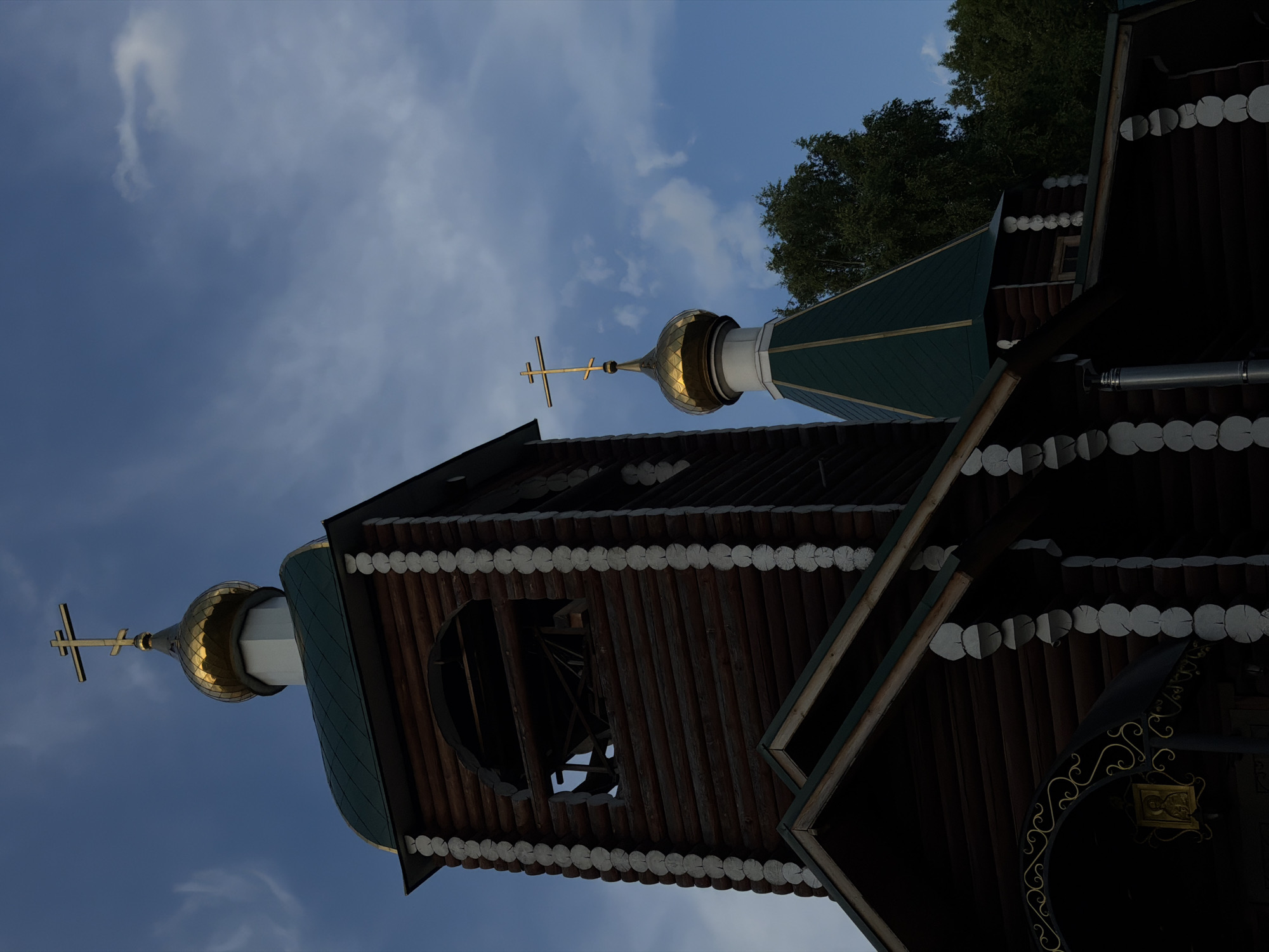 The image shows a wooden Orthodox church with green roofs and golden domes, set against a blue sky with scattered clouds. The church features a prominent bell tower with an arched opening and a bell visible within. The log construction of the building is decorated with white trim. A cross with three bars stands atop one of the golden domes. The foreground of the image is dominated by the dark wooden architecture of the church, with decorative gold scrollwork above an entranceway and a framed icon visible. Lush green trees are visible in the background on the right side of the image. The overall scene depicts a serene, religious structure in a natural setting. This church is likely the Church of the Ascension of the Lord, located in the city of Chaikovsky, Russia.