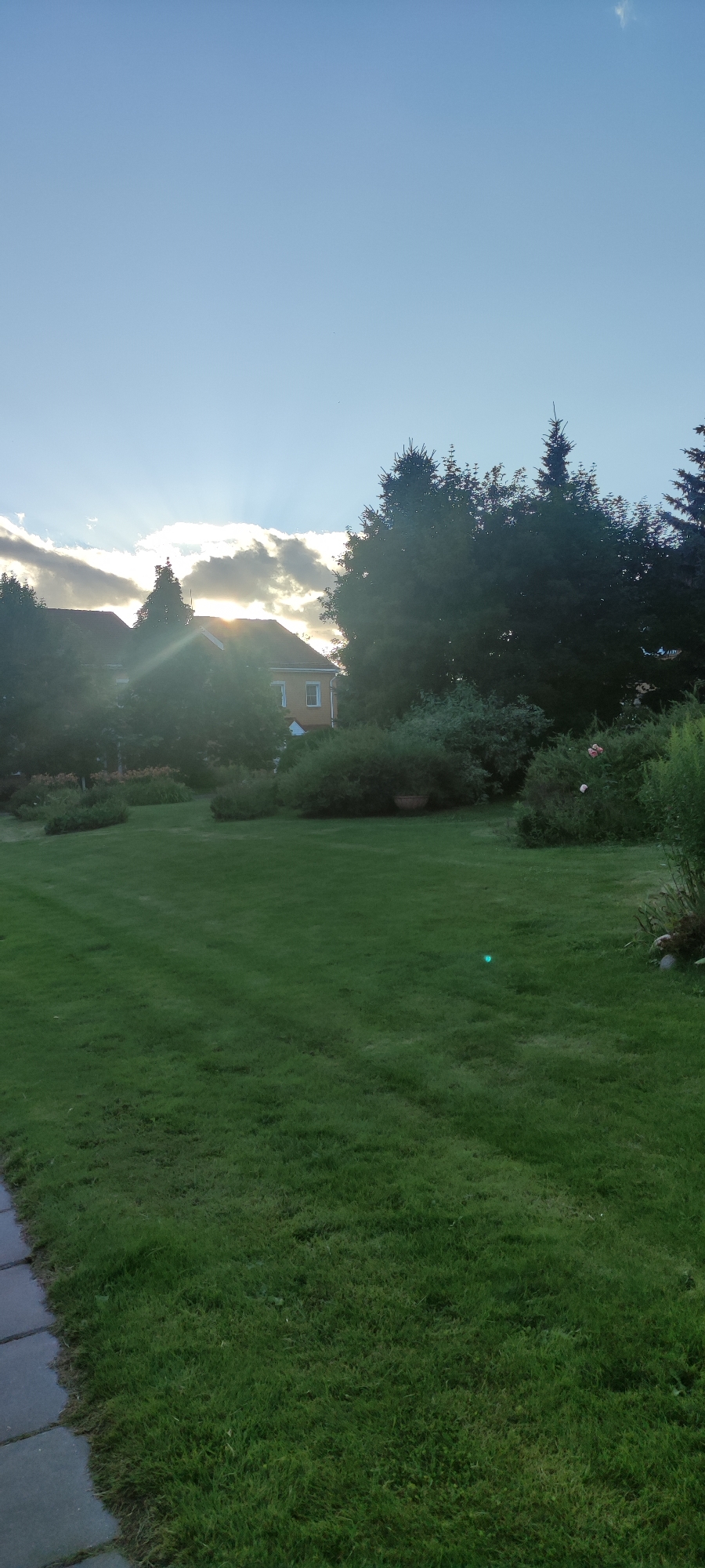 This is a ground-level, wide-angle photograph taken outdoors in Rosinka, Russia, capturing a suburban residential setting during the late afternoon or early evening. The scene is dominated by a well-maintained, lush green lawn that occupies the majority of the foreground and middle ground. A paved pathway, made of large, rectangular gray stones, runs along the left edge of the frame.
In the background, a two-story house with a reddish-brown roof and yellow siding is partially visible, nestled amongst dense, dark green trees and shrubs. The sun is setting behind a band of dramatic, dark clouds in the upper left portion of the sky, casting a warm, golden light that pierces through the clouds and creates sunbeams. This light also reflects off the windows of the house and illuminates the foliage. The sky above is a clear, pale blue, with a few wispy clouds.
The overall impression is one of a tranquil, domestic landscape bathed in the soft glow of a setting sun. There are no people or animals visible in the image, and the atmosphere is serene and quiet. The presence of the house, lawn, and landscaping suggests a residential area.