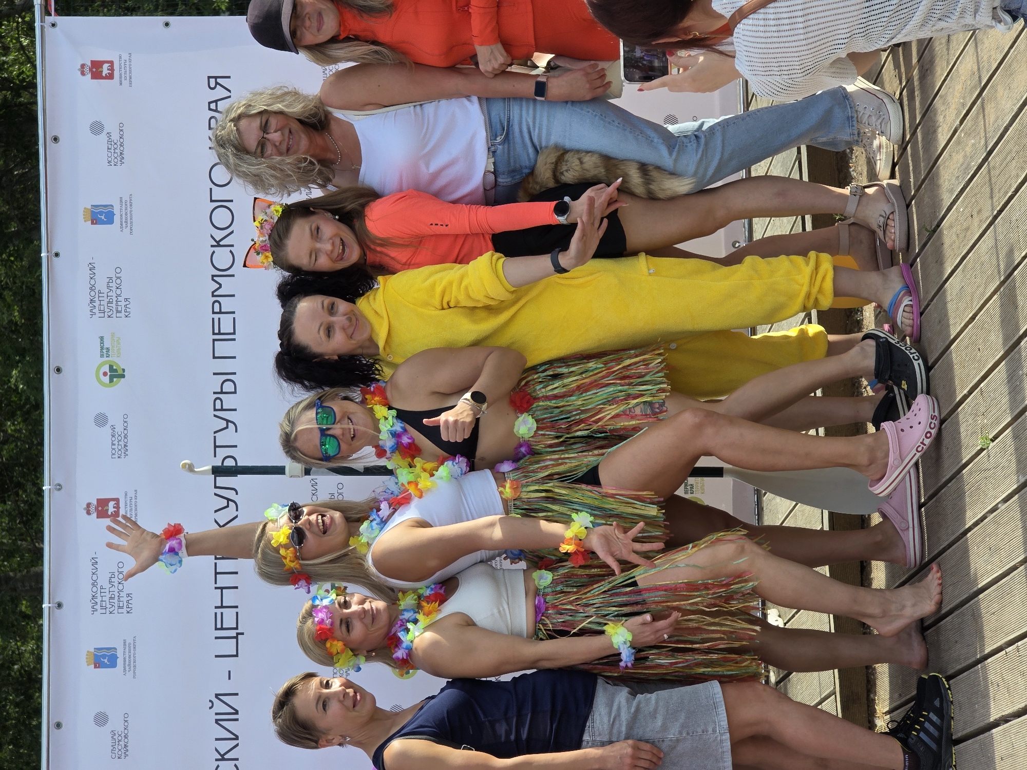 Eight women are posing for a photo in front of a banner. The banner has logos and text that appears to be in Russian. The women are wearing colorful, festive clothing, including lei necklaces and flower crowns. Several of them are wearing hula skirts. The ground is made of wooden planks. The women appear to be at an outdoor event. The banner indicates the event is associated with the Chaikovsky Center of Culture, Perm Krai.