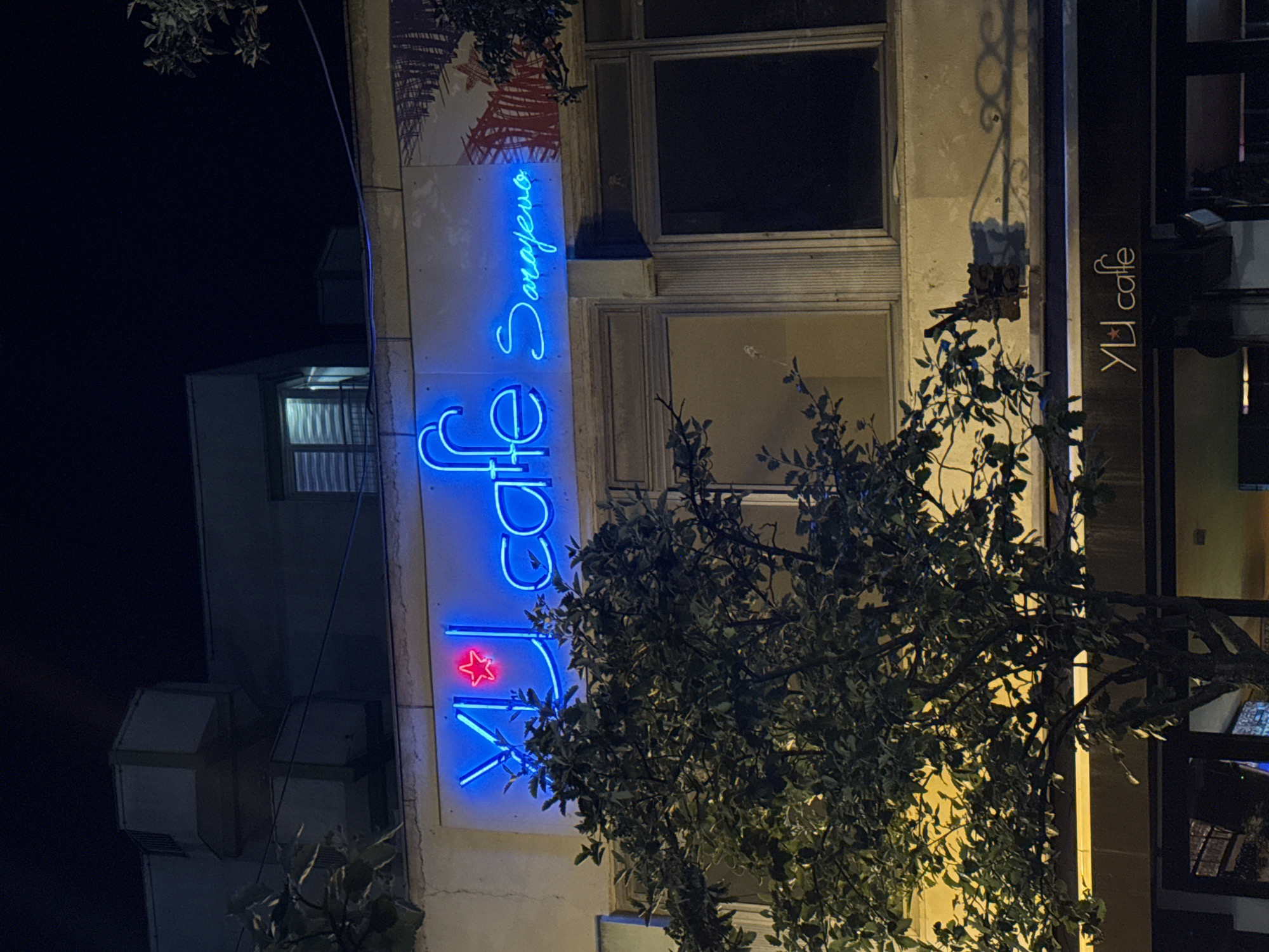 This image shows a neon sign for "YU cafe Sarajevo" on the facade of a building at night. The sign is blue and features a red star. A tree with dense green foliage partially obscures the sign. The building appears to be a commercial establishment, with windows visible on the upper floor. Below the neon sign, a darker awning displays the same "YU cafe" logo in white lettering. The overall environment is dark, suggesting nighttime, with artificial light illuminating the building and signage. The location is identified as Ilidža, Bosnia and Herzegovina.