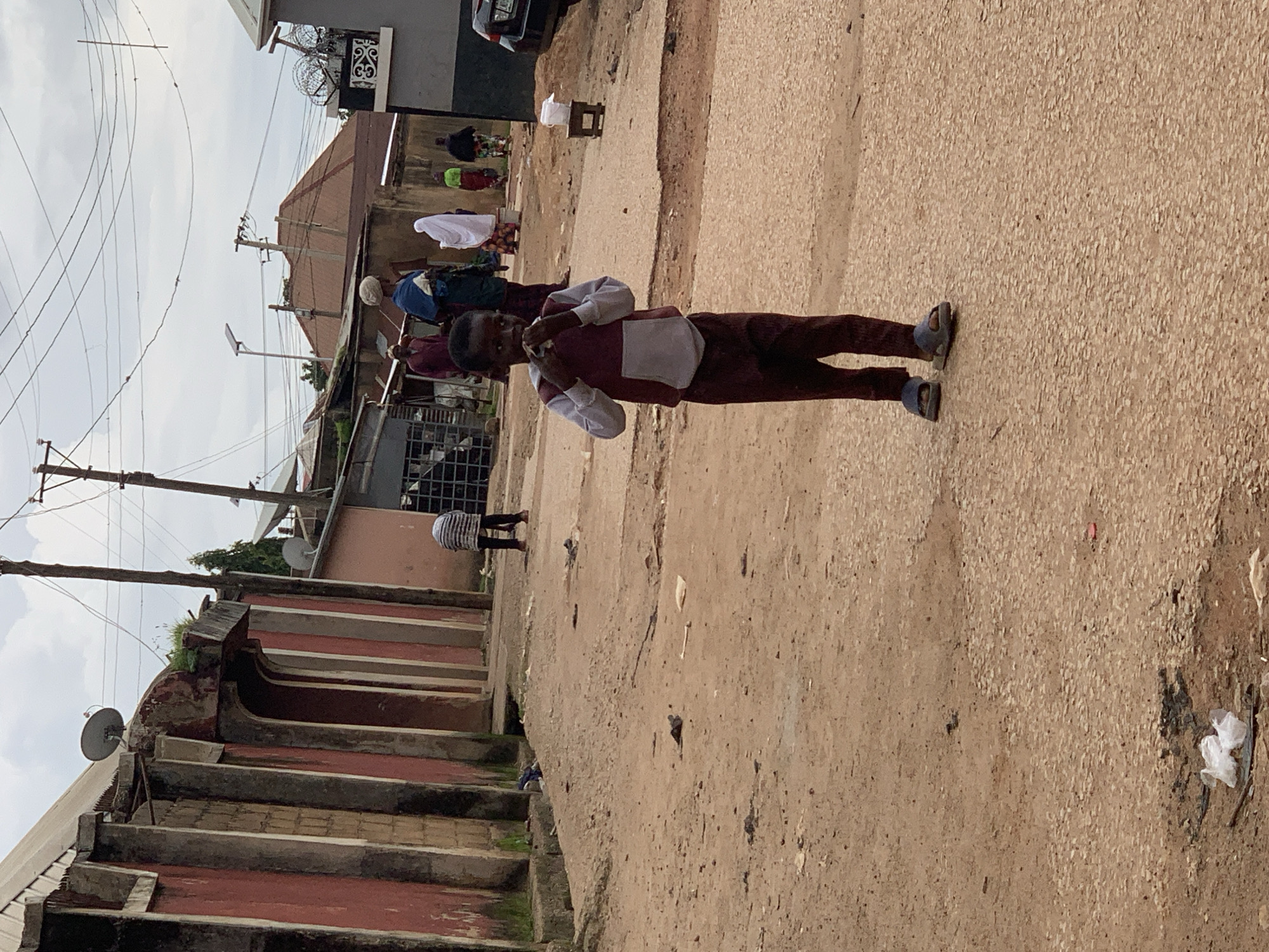 In a dusty street in Jos, Nigeria, a young boy stands in the foreground, holding his hands to his mouth. He is wearing a maroon and grey hooded sweatshirt with maroon pants and blue sandals. In the background, several adults are walking away from the camera. The street is unpaved and appears to be a residential area, with buildings lining one side. The sky is overcast, and there are utility poles and wires visible. There is debris scattered on the ground.