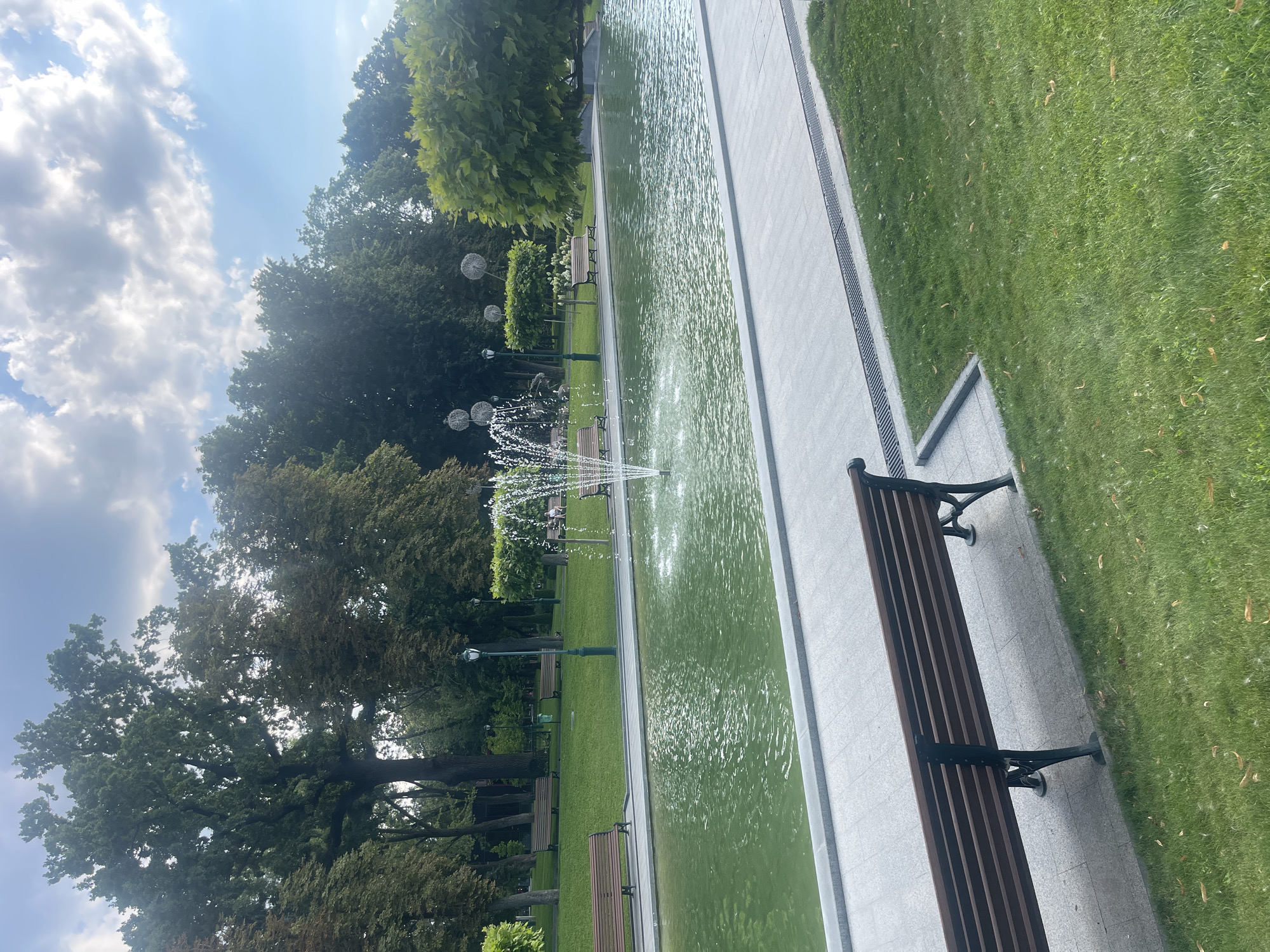 A tranquil park scene unfolds with a central water feature, a pond with a spouting fountain, reflecting the lush green surroundings. The pond is bordered by a paved walkway and a manicured lawn. Several park benches are strategically placed throughout the scene, offering spots for repose. Towering trees with dense foliage form a verdant backdrop, some adorned with whimsical, orb-like decorations. A cloudy sky with hints of blue completes the serene atmosphere. The image captures a peaceful moment in an urban park setting.
