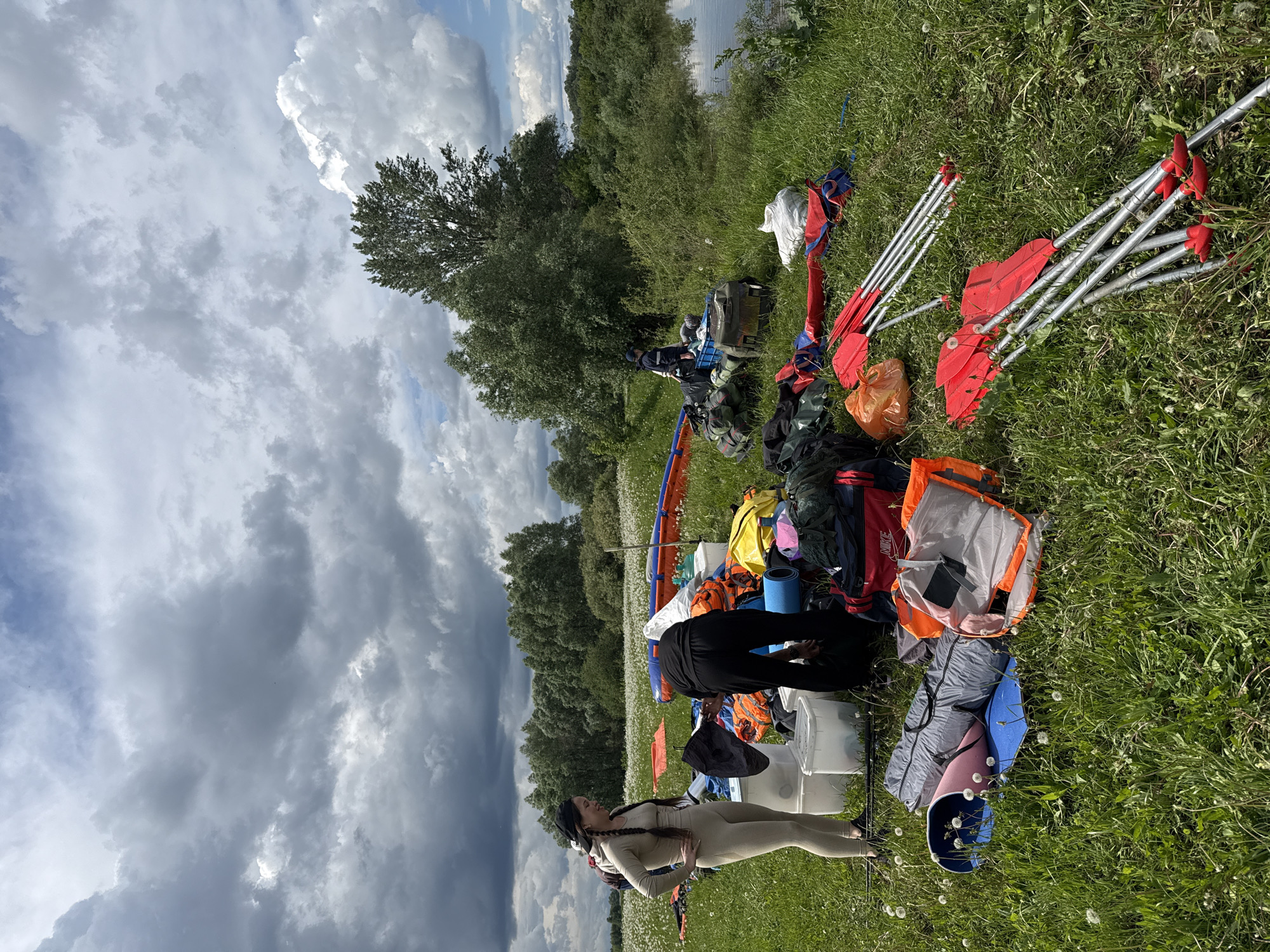 The image captures a grassy outdoor setting. Two individuals are present, one wearing a beige outfit and a head covering, standing with hands on hips. The other is partially visible, wearing black clothing and bending over a pile of gear. The gear includes backpacks, paddles with red blades, and various containers and camping equipment. The background shows trees and a cloudy sky. The location is unclear, Nazyar, Russia.