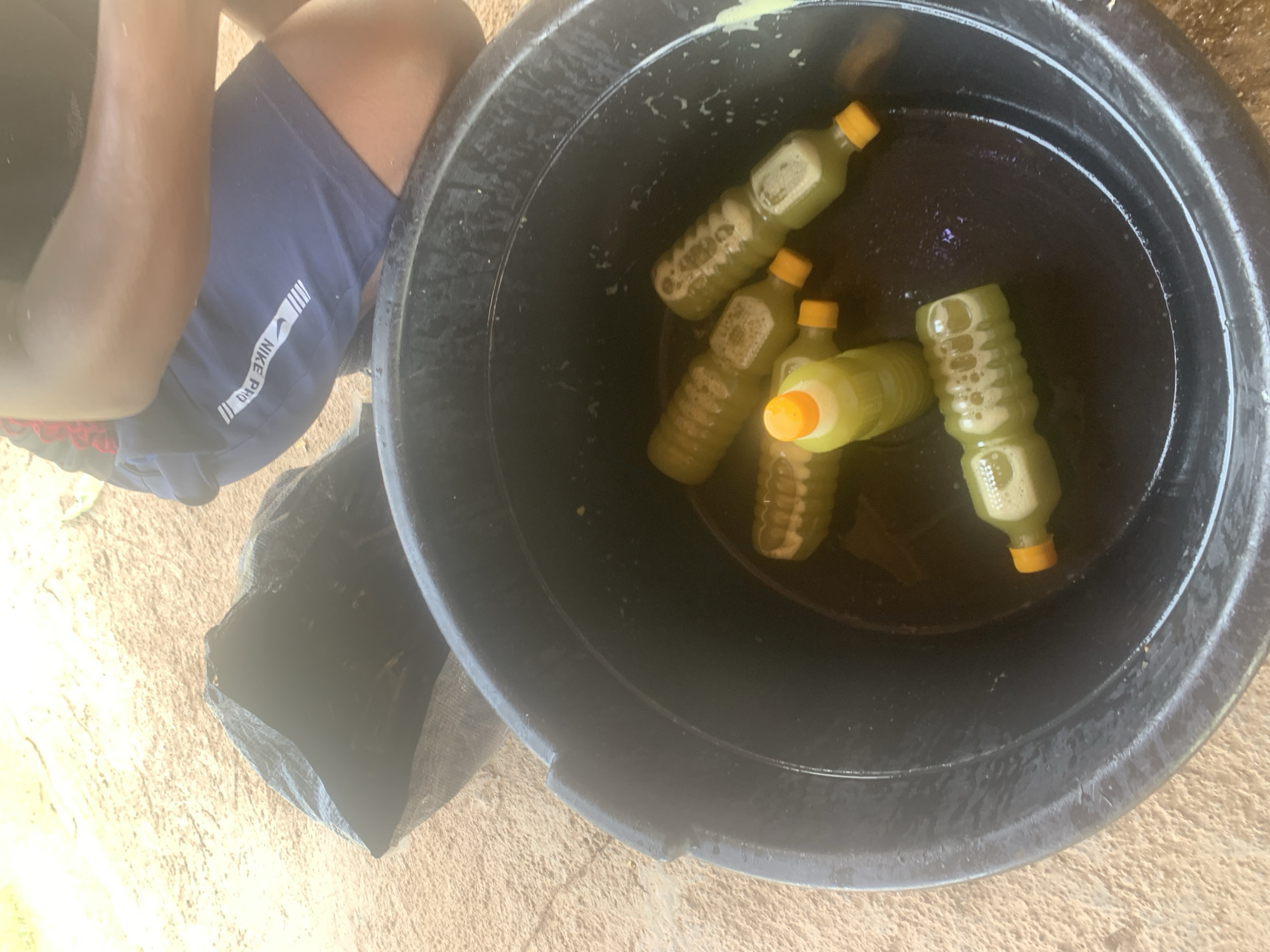 In a high-angle outdoor shot on the outskirts of Jos, Nigeria, a large black plastic basin is filled with murky liquid and several green plastic bottles with yellow caps. The bottles appear to contain a light green liquid with some foam. A black plastic bag lies to the left of the basin, partially obscuring the sandy ground. In the upper right corner, a person's legs are visible, wearing dark blue Nike Pro shorts. The overall impression is one of a food or beverage preparation setting.