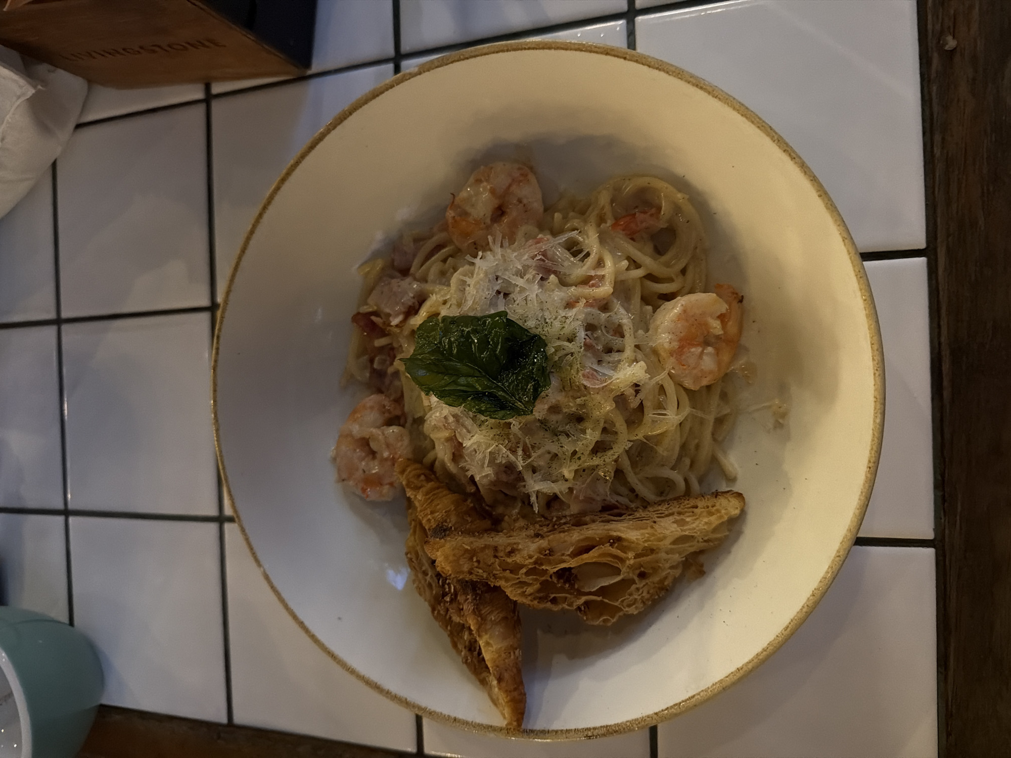 An overhead shot shows a white bowl of spaghetti with shrimp and toast on a white tiled surface. The spaghetti is coated in a creamy sauce and topped with shredded parmesan cheese and a green leaf. Two pieces of golden-brown toast, appearing to be cut croissants, are placed to the left of the spaghetti. The bowl has a light brown rim. In the upper left corner, a teal bowl is partially visible. To the right of the bowl, a brown paper bag with the embossed text "LIVINGSTONE" is visible. The background consists of white square tiles with dark grout lines. The scene appears to be an indoor setting, possibly a restaurant, given the presentation of the food and the tiled surface. The image location is North Kuta, Indonesia.