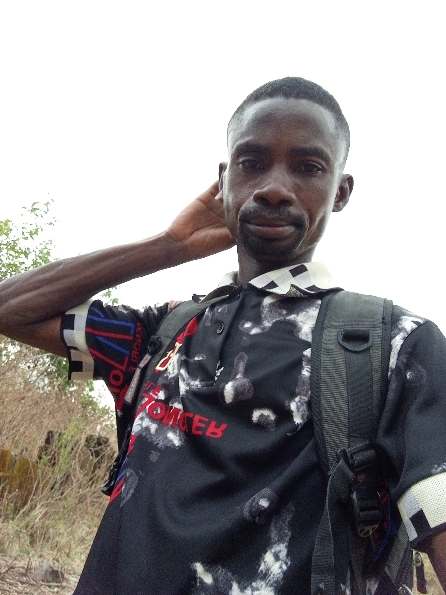An adult male stands outdoors, posing for a photo. He has dark skin, short hair, and a goatee. He's wearing a black shirt with a patterned collar and sleeves, the word "MOBILE" printed across the front. He is wearing a backpack. His left hand is raised behind his head. In the blurred background are bushes and vegetation, likely indicating a natural environment. The specific location in Lokoja, Nigeria, cannot be determined based solely on visual information.
