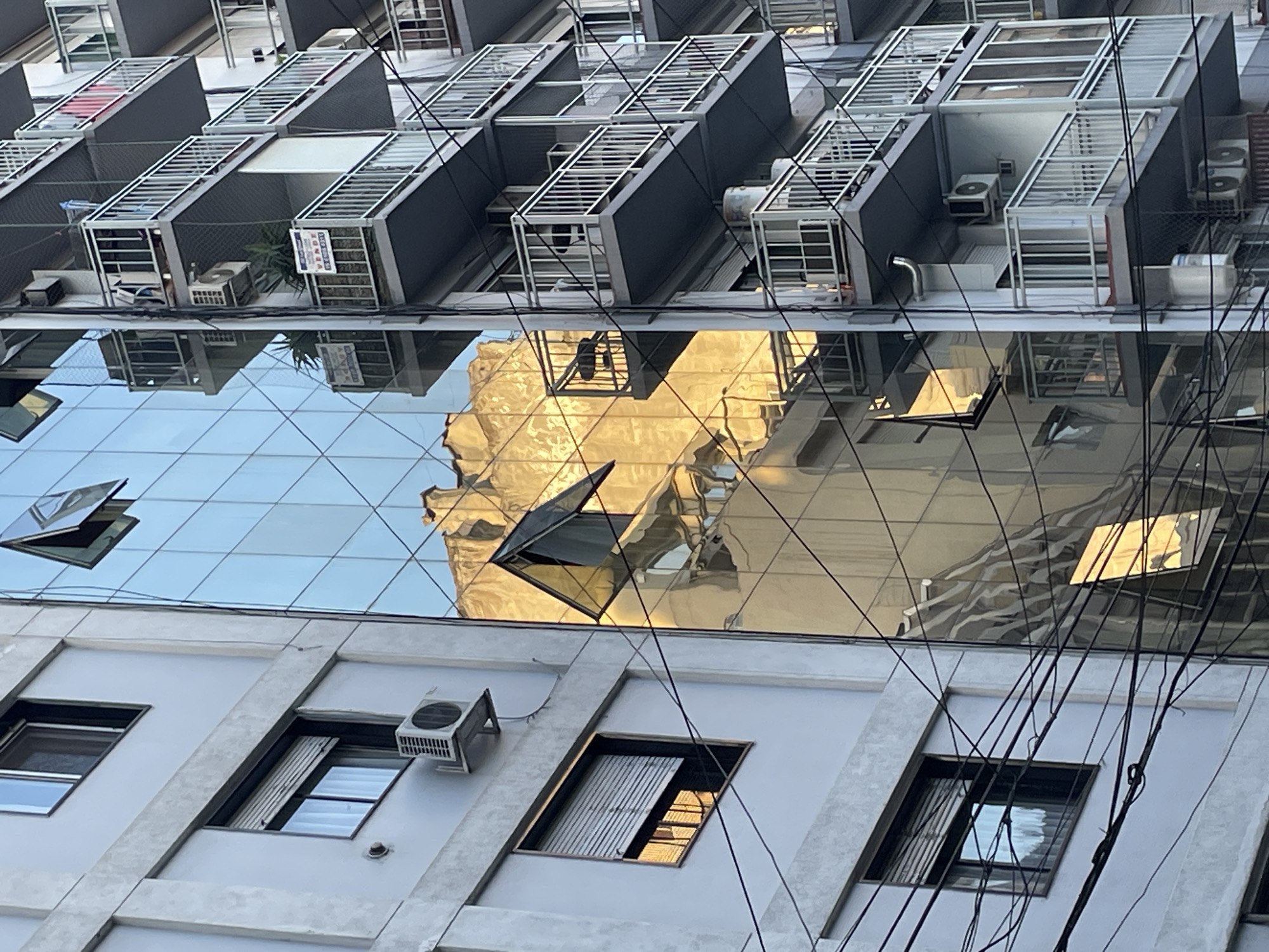 The image captures a scene in Buenos Aires, Argentina, presenting a multi-layered perspective of urban architecture. In the foreground, a building with light-colored concrete and rectangular windows, some partially open, features an external air conditioning unit. Above, a glass facade reflects a golden building, distorted by the facets of the glass. In the upper background, multiple cantilevered structures with metal grating, some housing air conditioning units, are visible. Numerous power lines crisscross the view, adding to the urban density. The lighting suggests either sunrise or sunset, contributing to the warm tones of the reflected building.