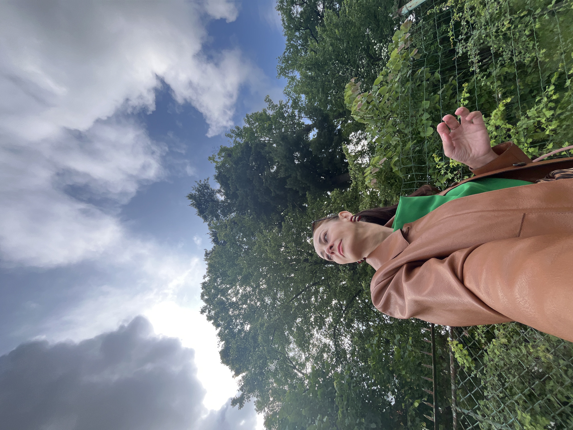 A woman is captured in a low-angle, selfie-style shot, looking up towards the sky. She is wearing a brown leather jacket over a green garment and has dark hair. Her facial expression suggests a moment of contemplation or observation as she gazes upwards. The background is a mix of lush green foliage from trees and a chain-link fence. Above, the sky is a dynamic canvas of blue with scattered white and grey clouds, hinting at a partly cloudy day. The lighting suggests it is daytime, likely late morning or afternoon, with some of the clouds appearing somewhat dark and heavy, possibly indicating recent or impending rain. The overall scene feels natural and outdoorsy, with the woman appearing to be in a park or garden setting. There is no visible text in the image.
