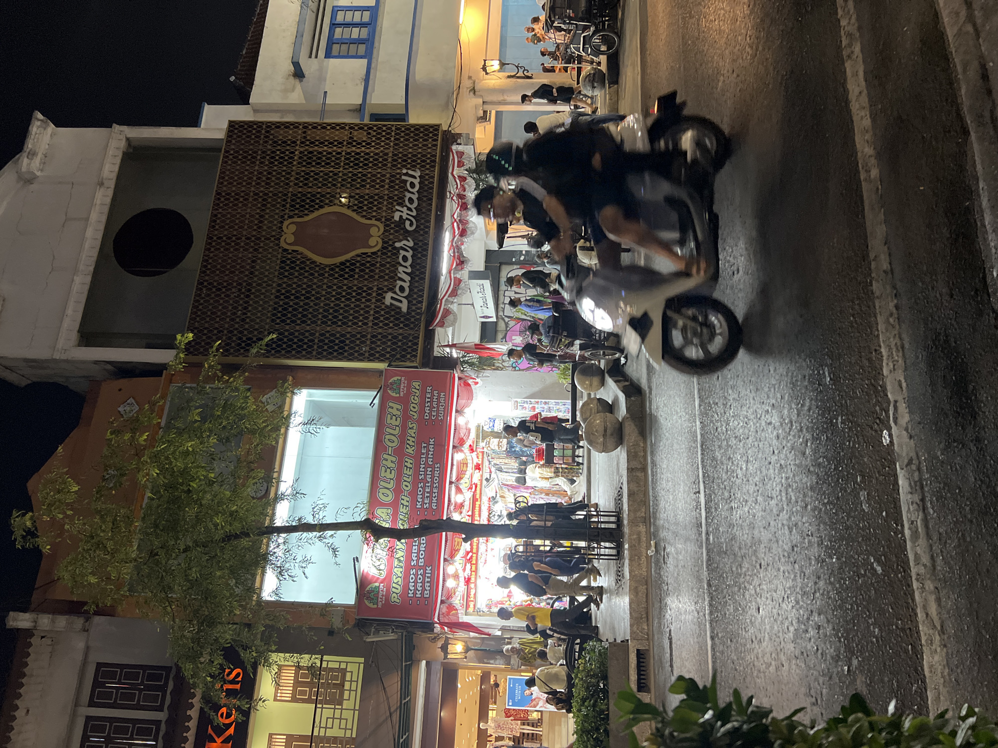 The image captures a night scene on Jalan Malioboro, a bustling street in Yogyakarta, Indonesia, known for its shops and vibrant atmosphere. The foreground is dominated by a wet asphalt road, reflecting the lights from the street and shops. A scooter carrying two passengers, a driver and a pillion rider, is in motion, appearing slightly blurred due to movement. The driver wears a black t-shirt and the pillion rider a black jacket and helmet.
On the right side of the road, a parked scooter-like vehicle with a passenger is visible. Further down the street, a group of people are standing and walking. In the midground, a street vendor's stall is open, adorned with red and white Indonesian flags, displaying various merchandise under bright lights. A medium-sized tree with sparse foliage stands in front of the stall. Several individuals are seen browsing or walking along the sidewalk.
The background features a row of multi-story buildings with distinct architectural styles. One building has a facade with a patterned screen and a decorative emblem. Another building displays a sign that reads "Danar Hadi." Above, the night sky is dark, indicating it is late evening or night. The overall scene conveys a sense of lively urban activity at night.