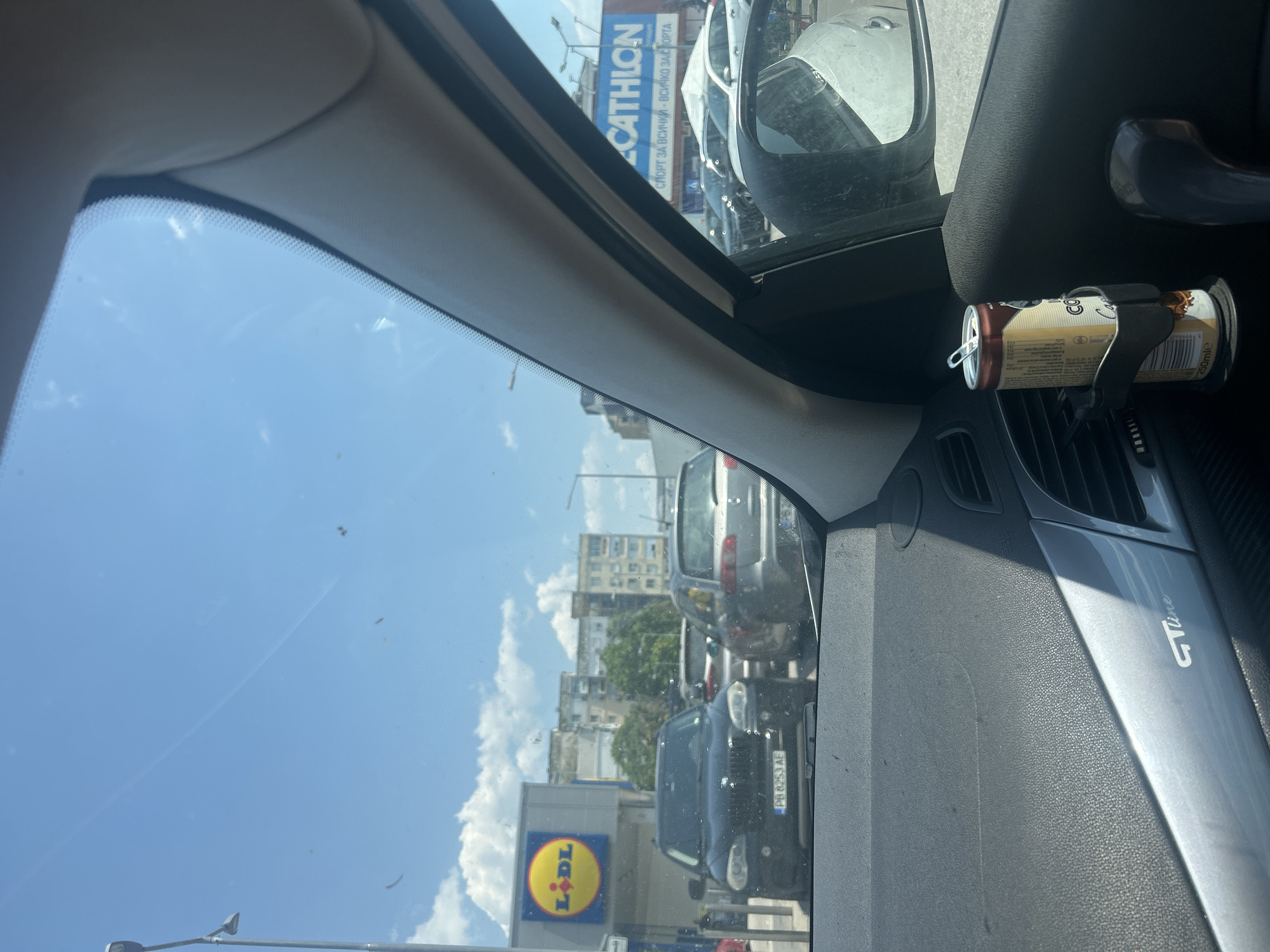 The image is taken from inside a car, looking out the windshield and side window. In the foreground, a can of "Coffee" drink is in a cup holder on the car's dashboard. The dashboard has a "GT Line" logo. Outside the car, several vehicles are parked in a line on a sunny day. To the left, a sign for "LIDL" is visible, indicating the car is likely in a parking lot near the supermarket. Further in the background, residential buildings are visible, and a large blue sign for "DECATHLON" with Bulgarian text translates to "SPORT FOR ALL - EVERYTHING FOR SPORT". The setting appears to be an urban environment in Plovdiv, Bulgaria. No people are clearly visible within the frame.