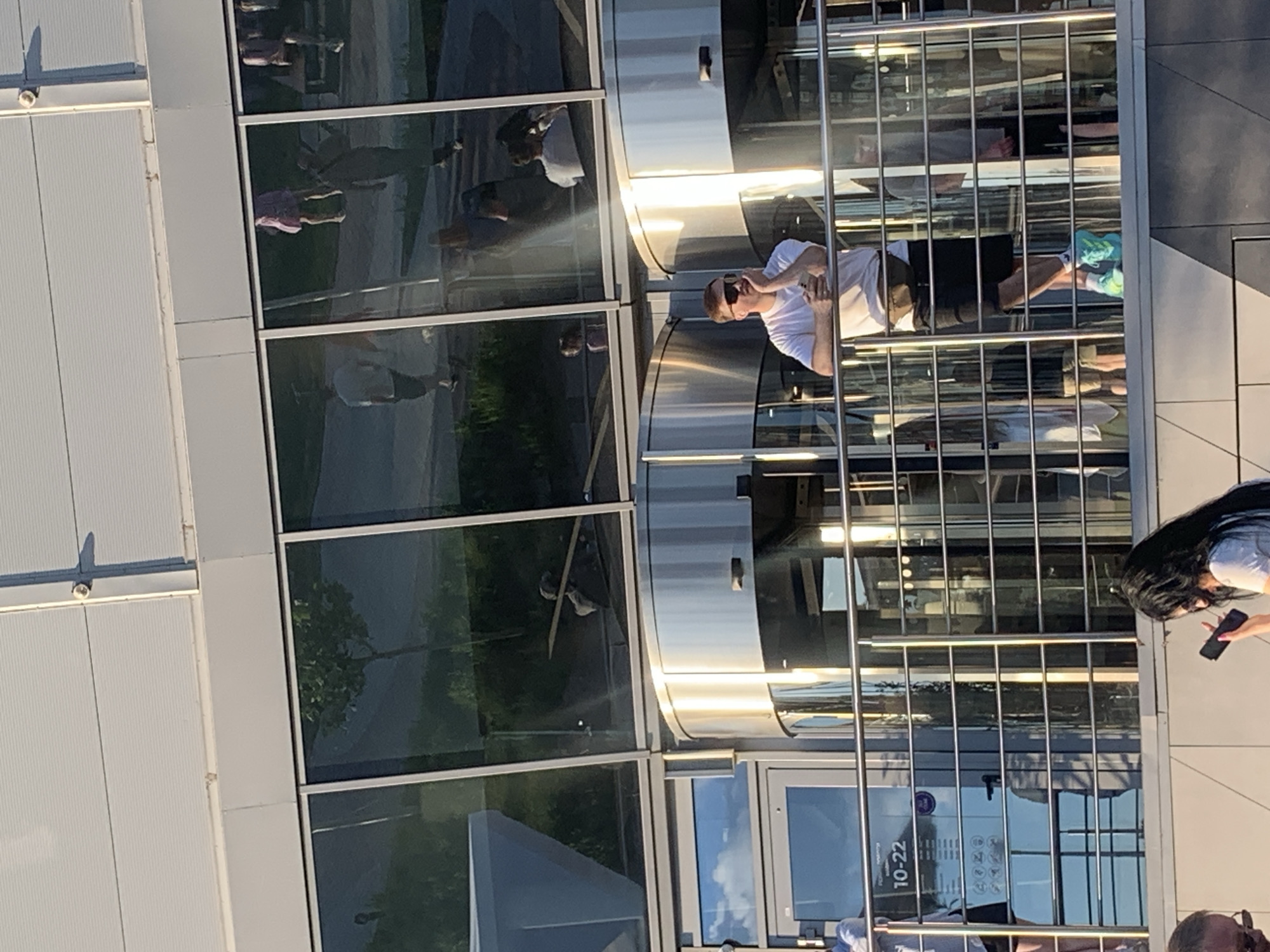 The image shows a group of people on an elevated outdoor walkway in front of a modern building with large glass windows. In the foreground, a man in a white t-shirt and black shorts is leaning on a railing, holding a phone and wearing sunglasses. He has a fanny pack around his waist and bright turquoise sneakers. Behind him, reflections in the glass show other people, including a woman in a pink dress. To the left, a sign on a glass door indicates operating hours from 10:00 to 22:00. The overall scene suggests a public space, possibly a shopping center or entertainment venue, given the revolving doors and the presence of multiple individuals. The lighting suggests it is daytime, with sunlight casting reflections. The specific location in Kyiv, Ukraine cannot be determined from the visual information alone, but the presence of cyrillic text on the sign could indicate a Ukrainian context.