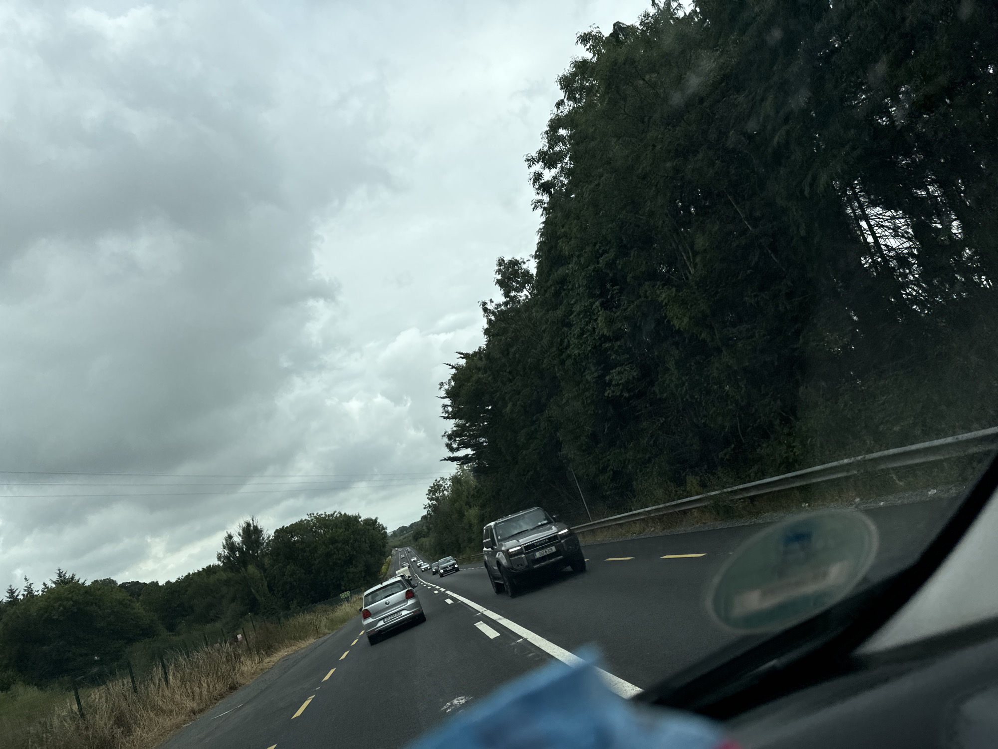 A grey, overcast sky looms over a rural road in Scartnamore, Ireland. The road, marked with yellow lines, curves slightly to the right. Several vehicles are traveling on the road, including a silver Volkswagen Polo in the foreground on the left, a dark SUV with a license plate reading "252-D-379" in the right lane, and other cars further ahead. Dense trees line the right side of the road, with a guardrail separating the road from the trees. The left side of the road features a grassy verge and more trees in the distance. The overall impression is of a slightly dreary day on a country road.