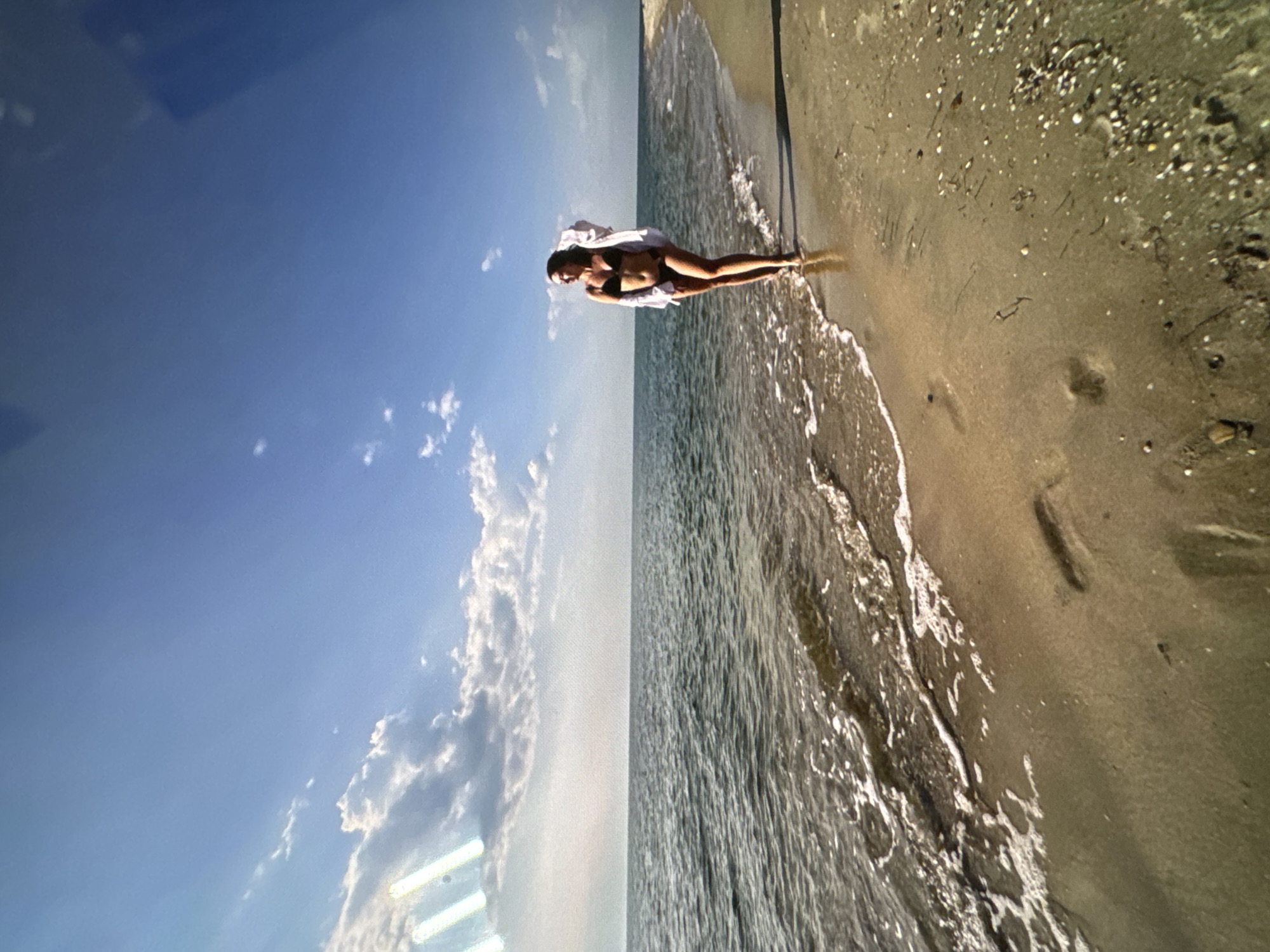A woman walks along a sandy beach with waves lapping at her feet. She is wearing a black bikini and a white open shirt. The ocean stretches out to the horizon under a bright blue sky dotted with fluffy white clouds. The beach is mostly sand with some scattered shells and debris. The scene appears to be a serene and sunny day at the coast.