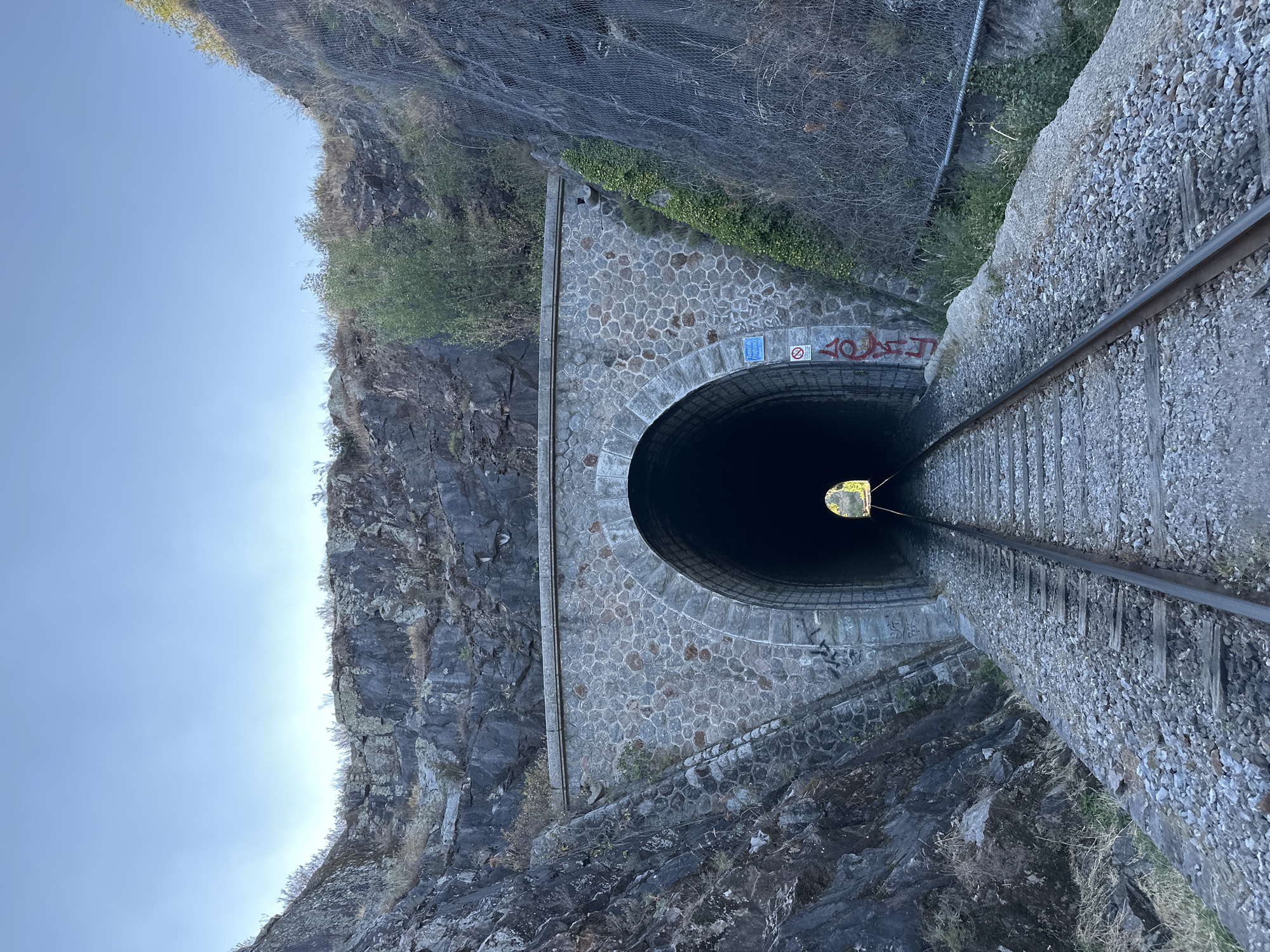 The image shows a view of a stone-arched railway tunnel entrance, a common sight on railway lines in the region, potentially near Générargues, France. The tunnel is set within a rocky, overgrown hillside. Railway tracks run from the foreground towards the dark opening of the tunnel, disappearing into its depths. The tunnel's exterior is constructed of rough-hewn stones, with some ivy growing on the right side. There is graffiti on the tunnel's facade, including red spray-painted lettering and a blue sign with white text. The sky above is clear and blue. The overall scene suggests a remote or rural location, characteristic of the French countryside.