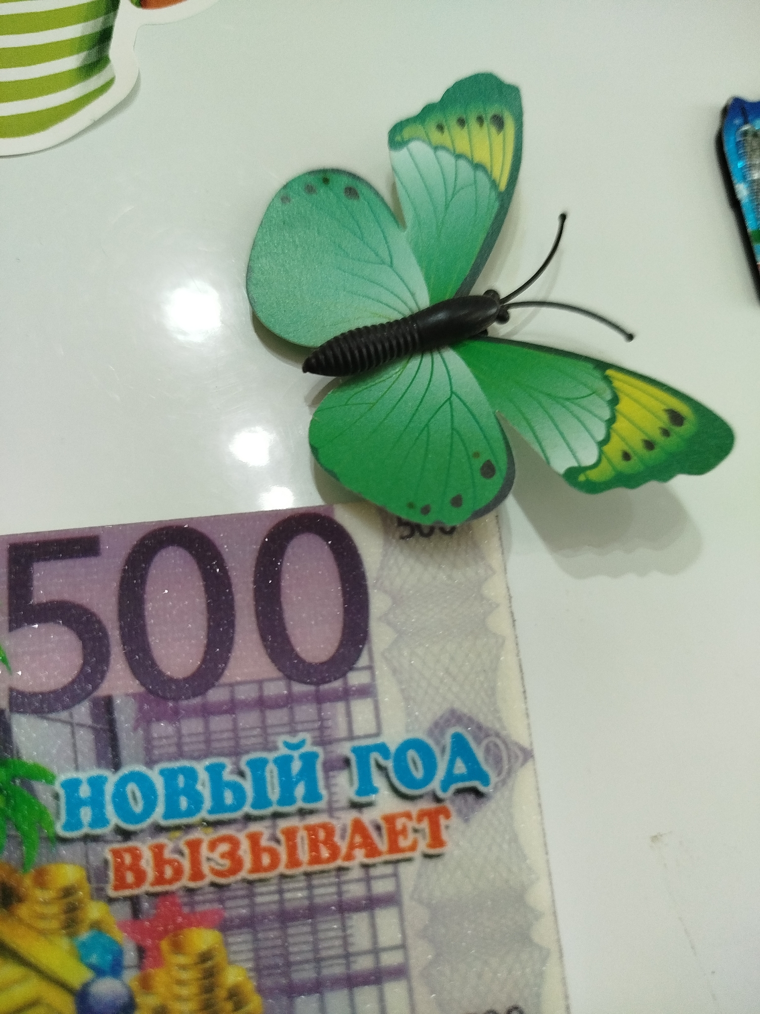 A green butterfly decoration is positioned on a white surface, partially obscuring a section of a banknote with the number "500" prominently displayed. Below the "500" are two lines of text in Russian: "НОВЫЙ ГОД" (New Year) in blue letters and "ВЫЗЫВАЕТ" (Causes/Evokes) in orange letters. The banknote itself appears to be a stylized representation, possibly a souvenir or decoration, with a purple hue and shimmering particles. To the left of the banknote, a hint of greenery, resembling palm fronds, is visible. The overall scene is a still-life arrangement of decorative items on a flat, light-colored surface. There is no indication of any specific location in Крывасельскі сельскі Савет, Belarus.