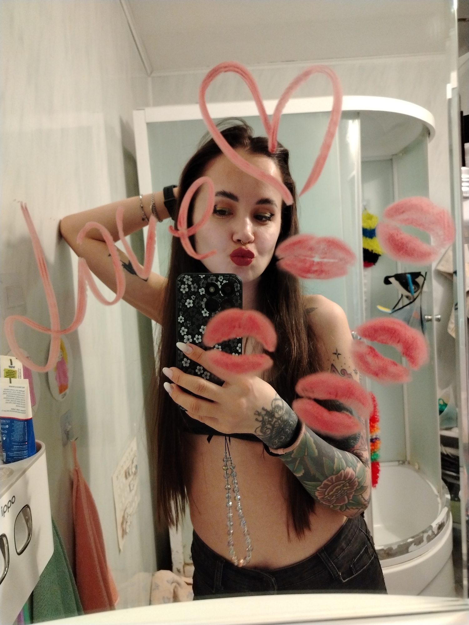 A woman is taking a selfie in a bathroom mirror. The mirror has pink lipstick marks and the word "Love" written on it. She is holding a black smartphone with a floral case. The woman has long brown hair, dark eyes, and a septum piercing. She is wearing a black bra and jeans. Her left arm is heavily tattooed with floral and geometric designs. In the background, there is a shower stall with various items inside and a sink area to the left. The bathroom appears to be in a residential setting. There is no evidence to support the location of this image being in Noviy, Russia.