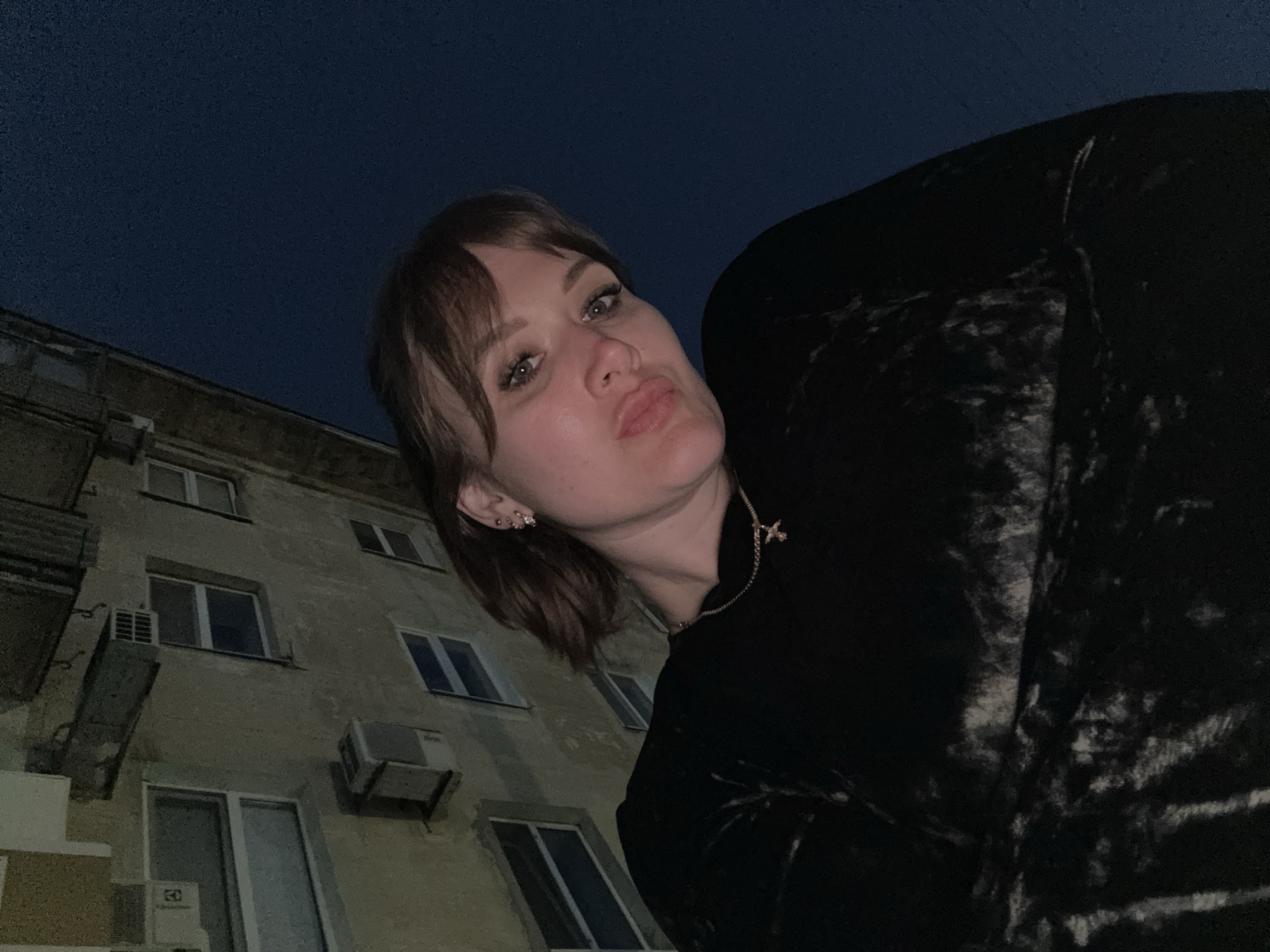 A low-angle shot shows a woman in Yevpatoriya, Ukraine, standing outside at night. The sky is dark blue. She is wearing a black velvet top and a necklace with a cross pendant. Her hair is brown and styled with bangs. She is looking down at the camera with pursed lips. Behind her is a beige building with several windows and air conditioning units.
